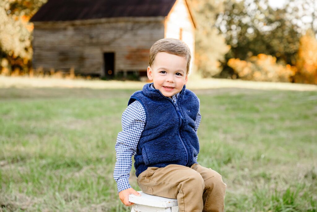 little boy dressed in a vest and pants sitting on a stool smiling with a fall barn behind him