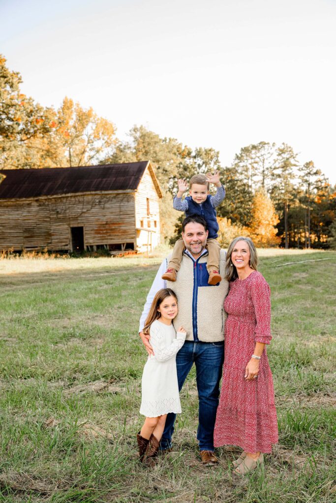 Full Family of 4 posed in front of a barn with youngest child on dad's shoulders raising his arms in excitement.