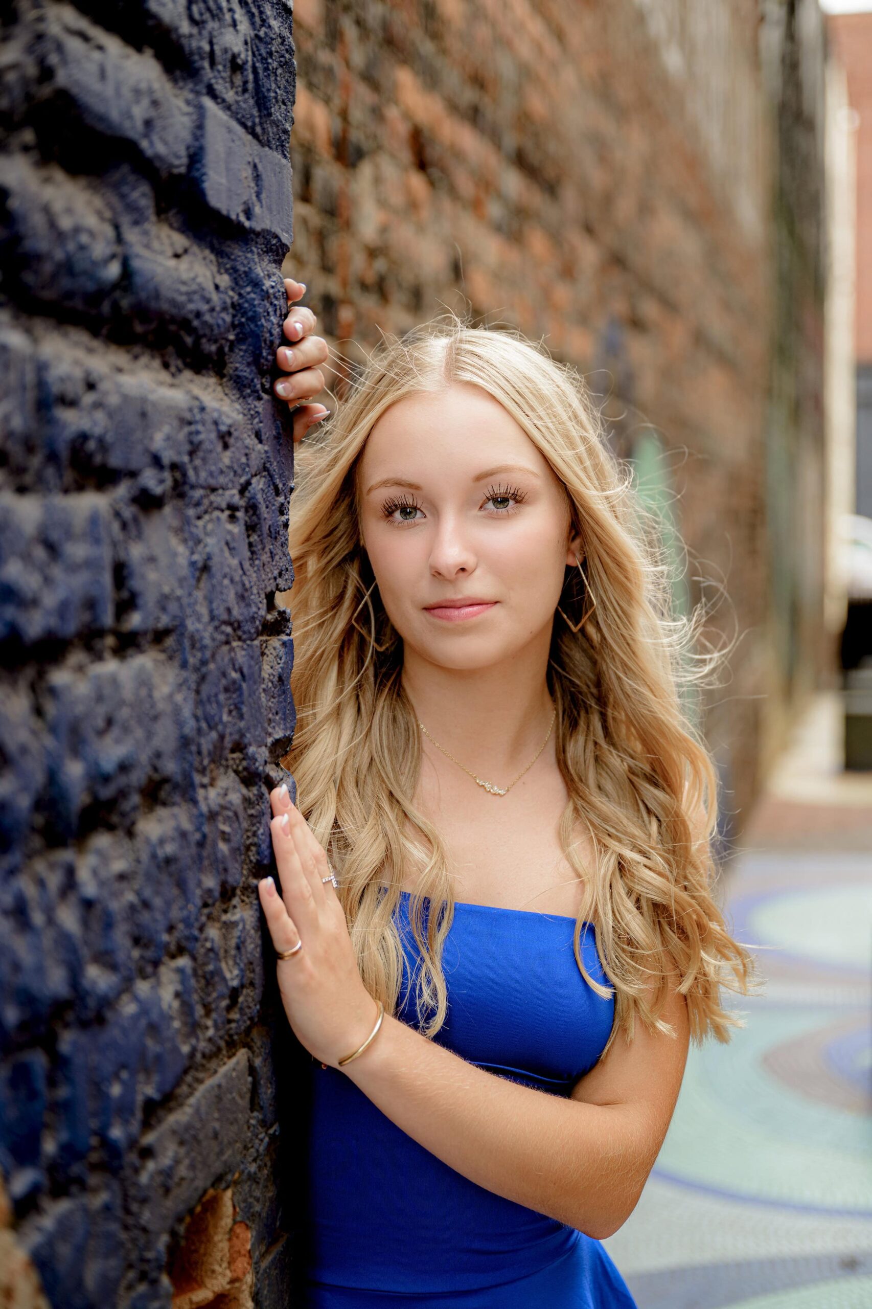 blonde senior poses with two hands on a colorful wall wearing a blue dress with her hair down