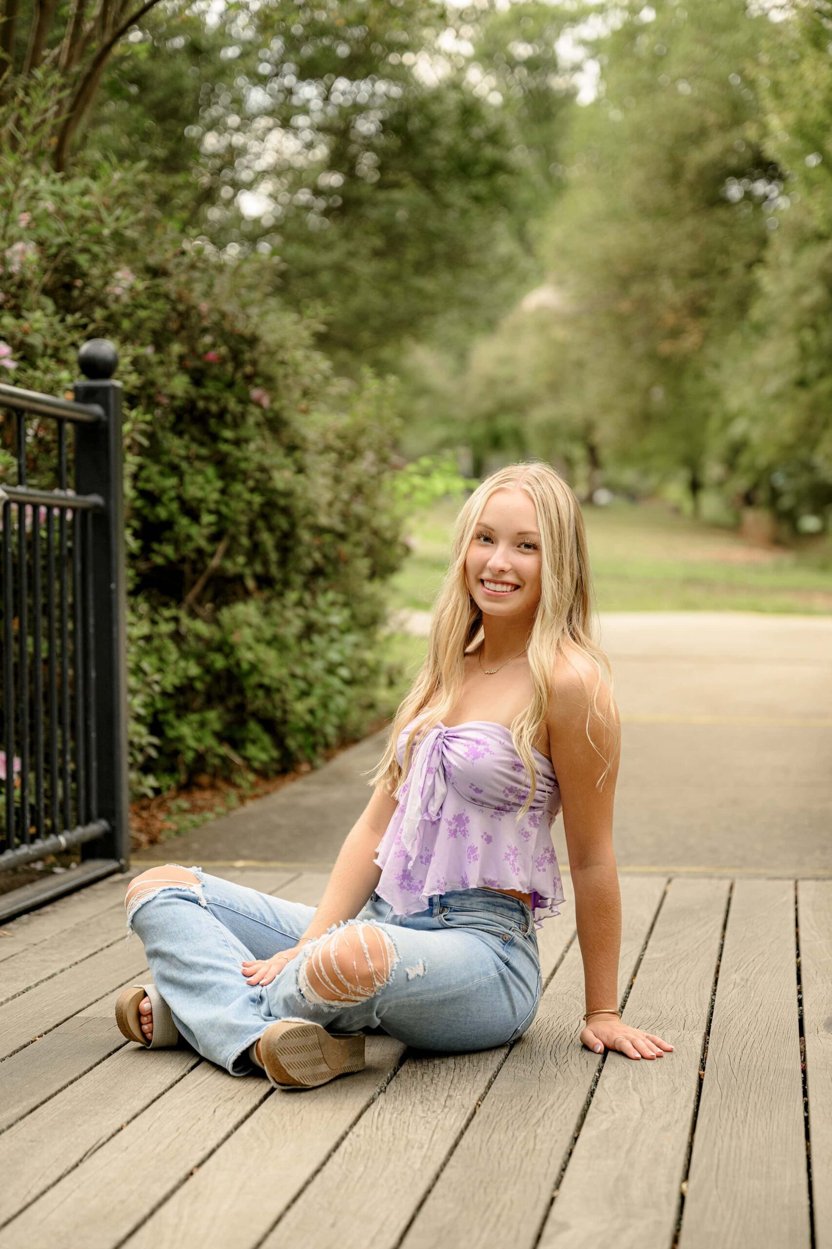 blonde senior sitting in a purple shirt and jeans in a rock hill garden on a bridge