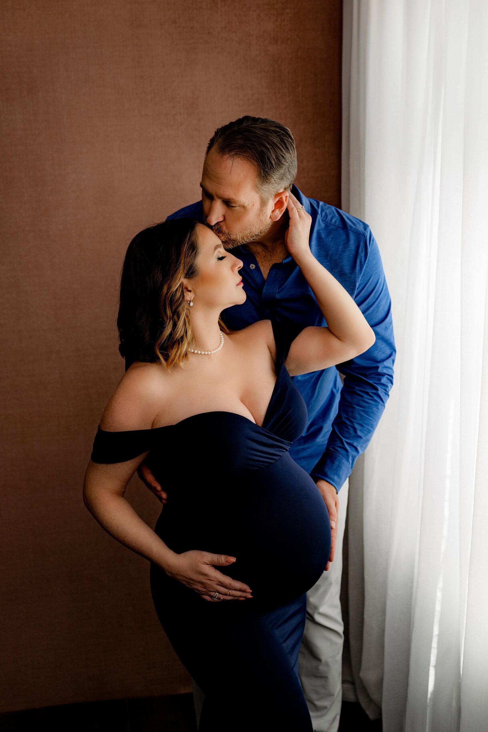 charlotte area couple poses together during their maternity session. mom and dad are both touching moms belly while dad leans over kissing her forehead. Mom to be is wearing a navy maternity client closet dress and dad is wearing a blue button down.