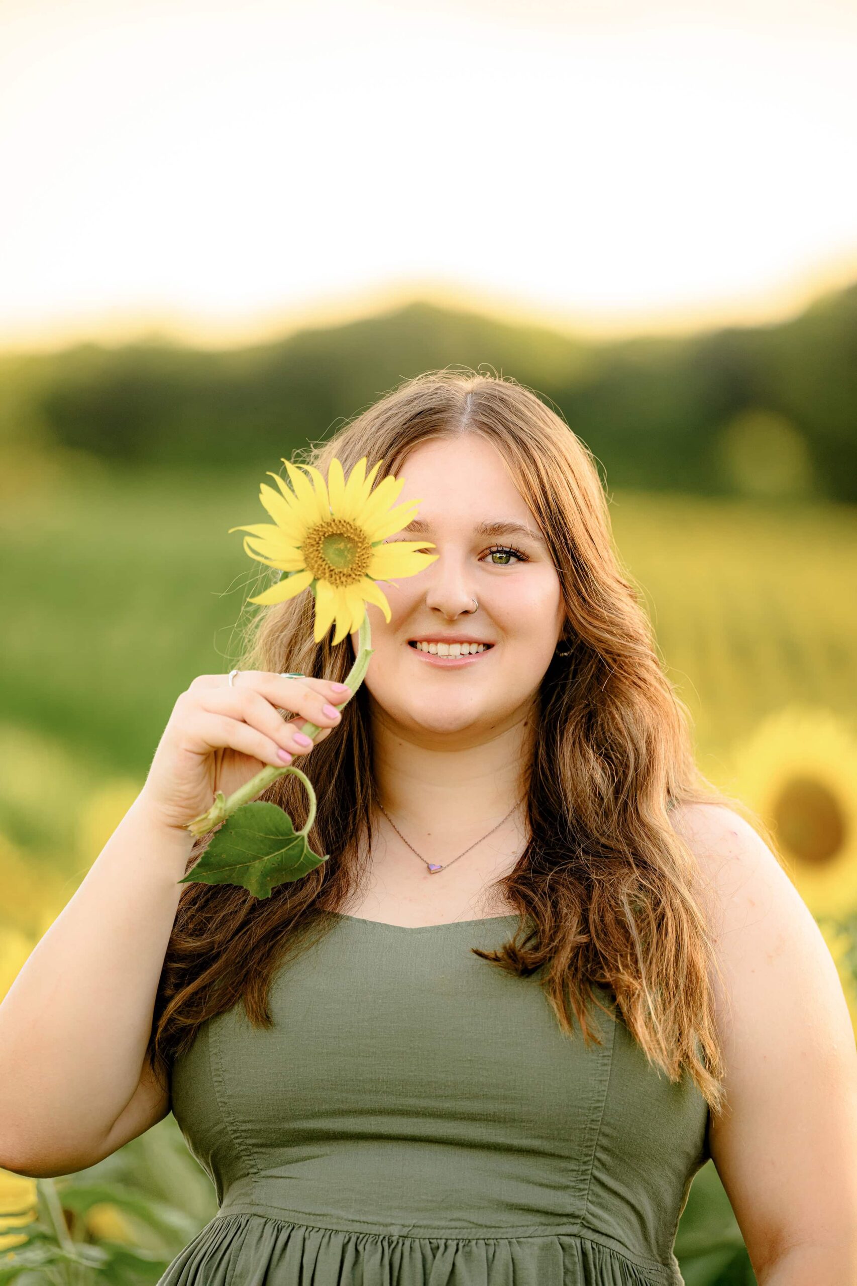 Cuthbertson senior with long dark hair holding a sunflower over her eye standing in a field in a green dress