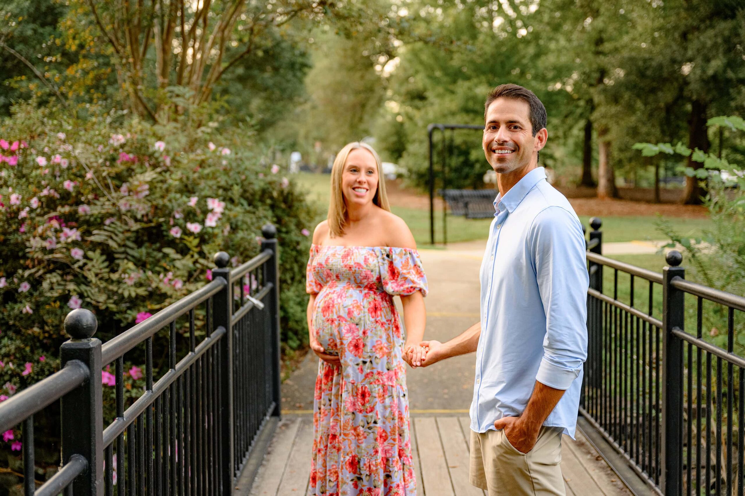 hand in hand expecting couple maternity image with mother in a floral dressed looking at her husband in a charlotte area garden during their outdoor maternity photography session