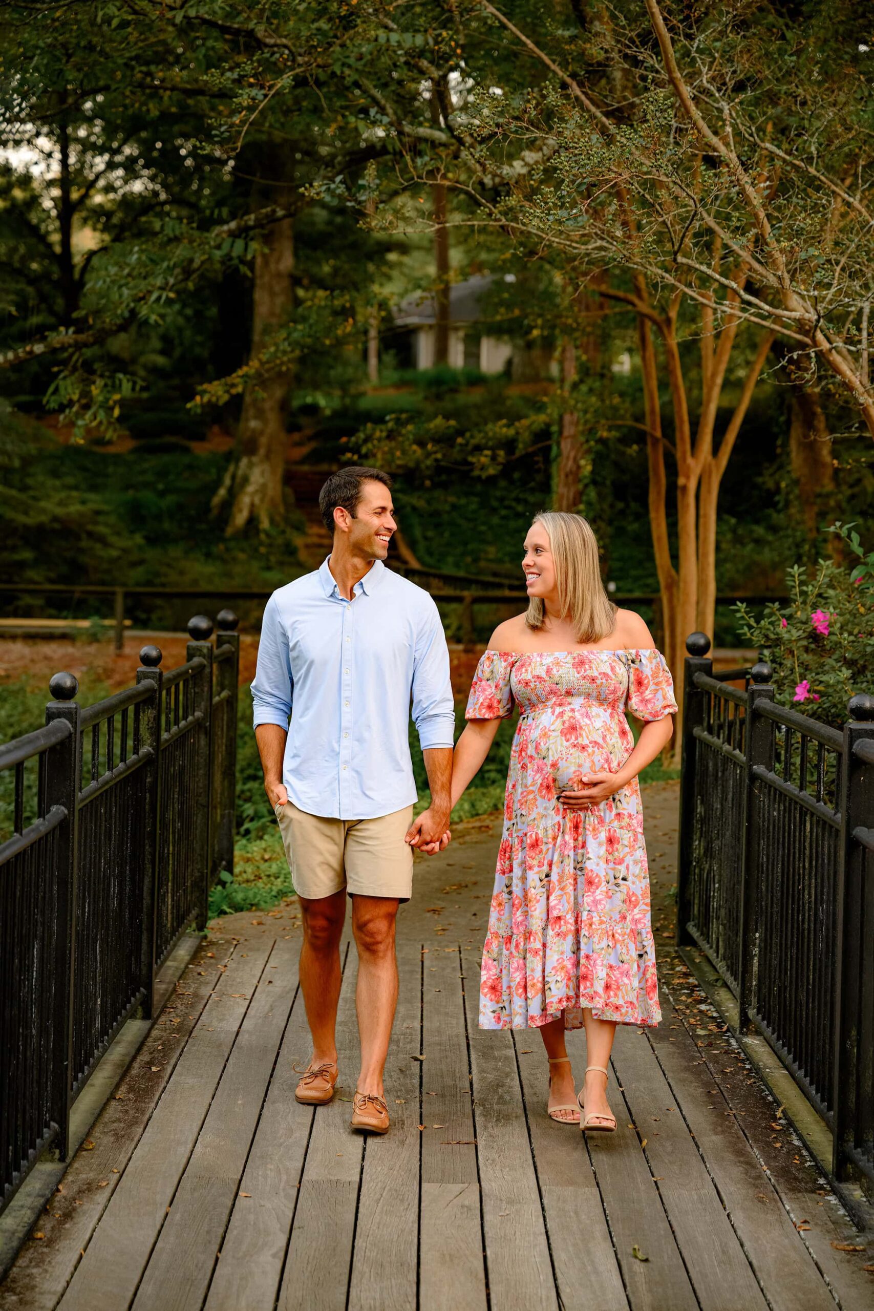 expectant couple stroll hand in hand during their outdoor maternity photography session with mom dressed in floral dress and dad dressed in blue button down and shorts