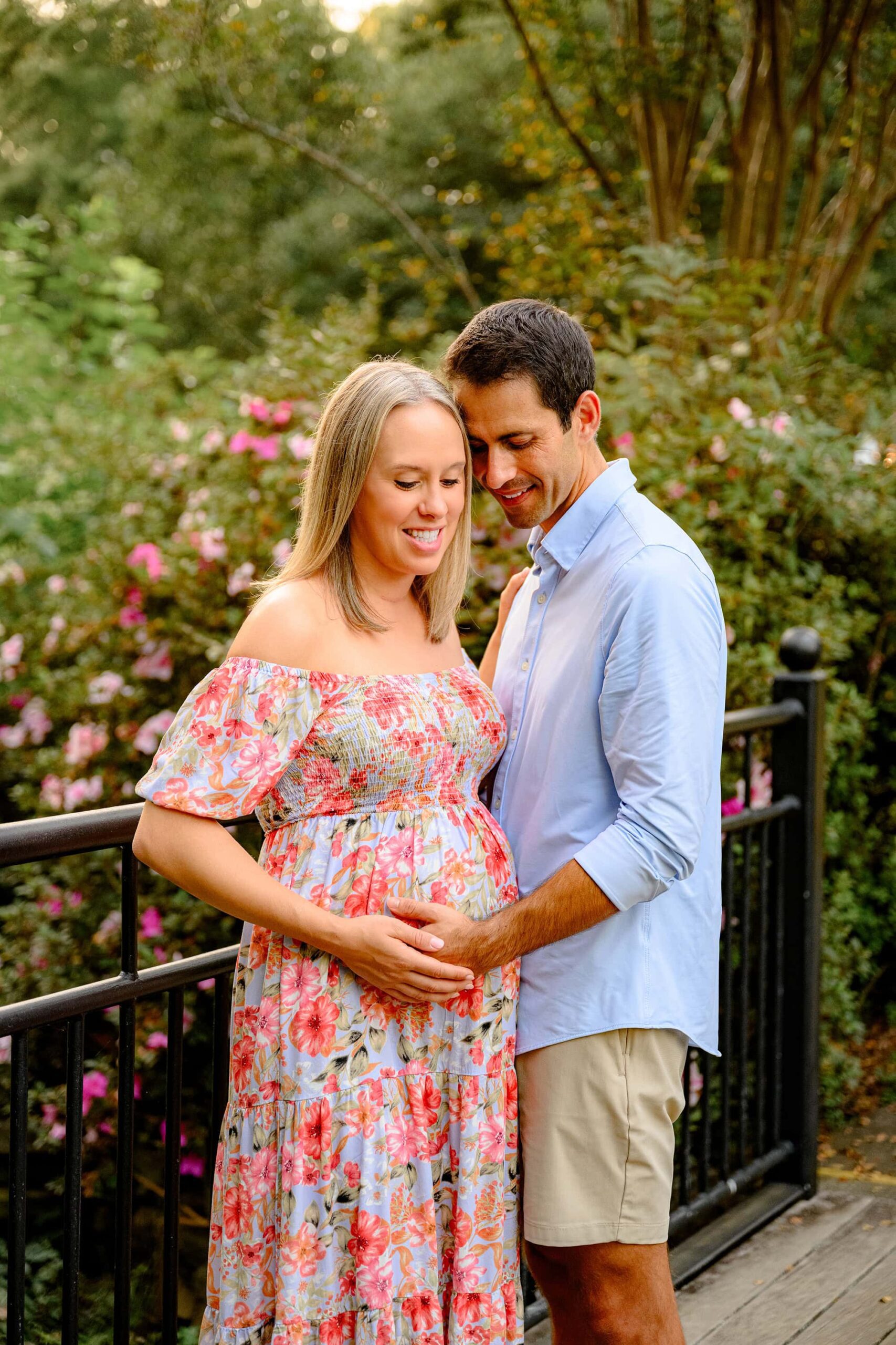 during their outdoor garden session an expectant mother in a floral dress and father dressed in blue button down while both looking at her baby bump