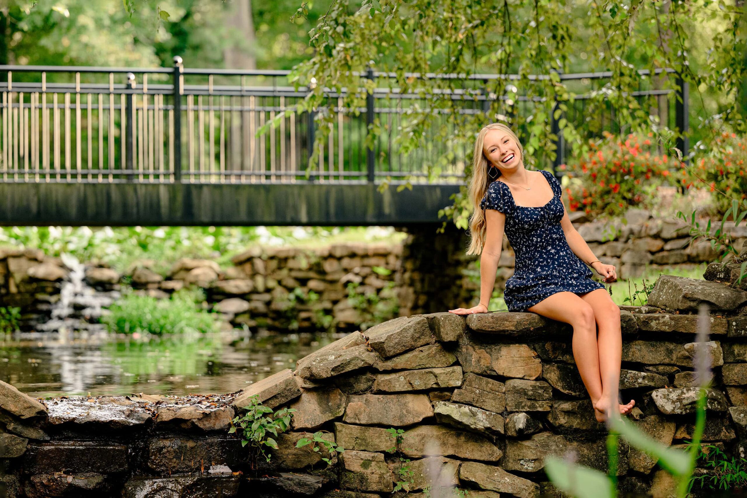 CHS senior girl dressed in a navy floral romper posed on a rock wall leaning over and laughing during her essential experience portrait session