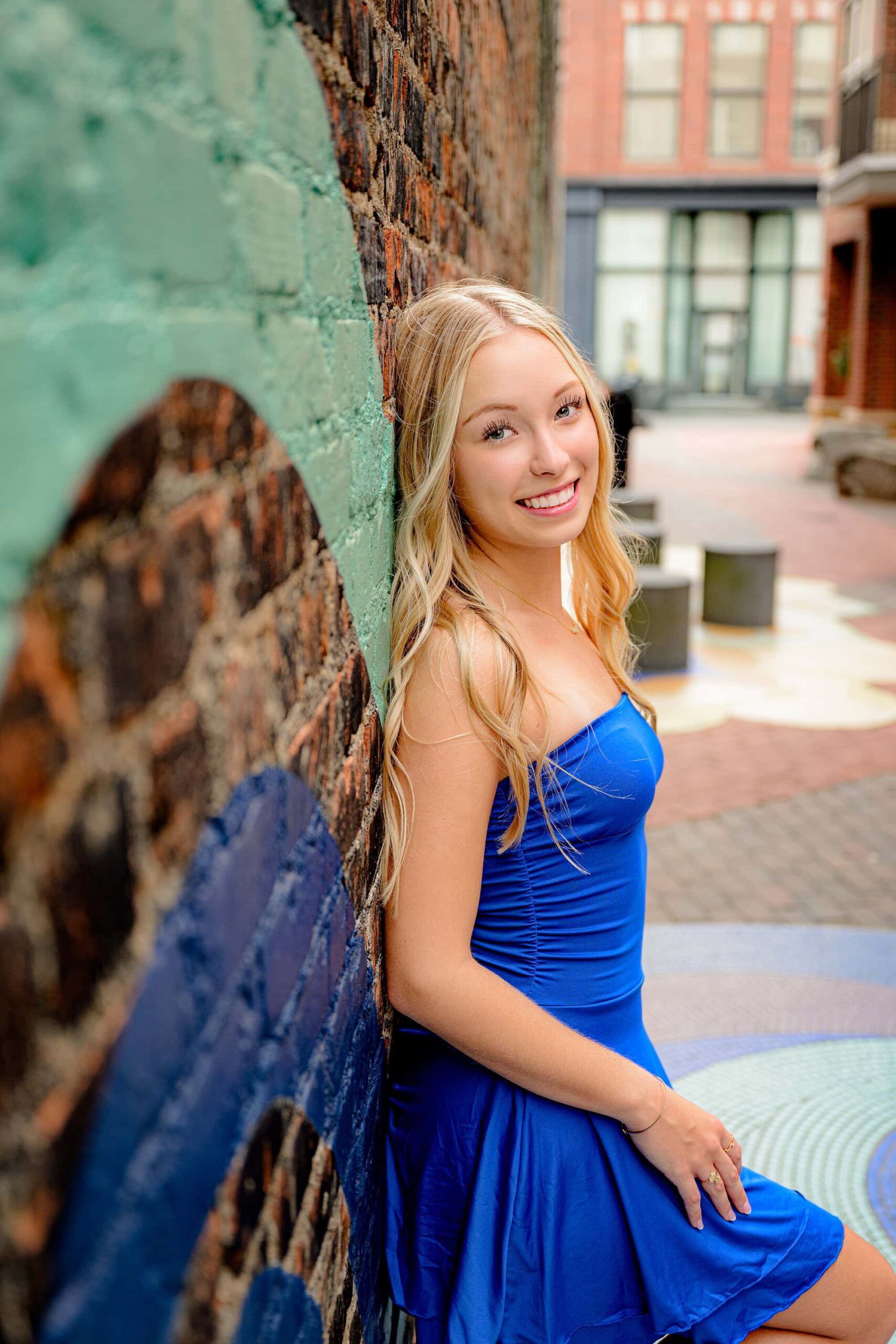 blonde senior girl dressed in royal blue tube dress sitting on a pedestal next to a graffiti wall wearing gold shoes during her essential experience grad session in rock hill