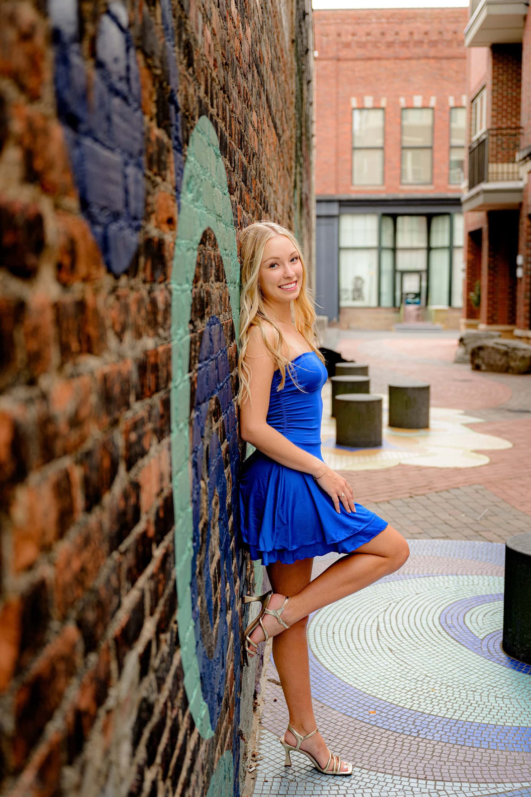 Blonde senior girl dressed in a royal blue tube dress, leaning against a graffiti wall and wearing gold shoes during her Essential Experience Grad Session.