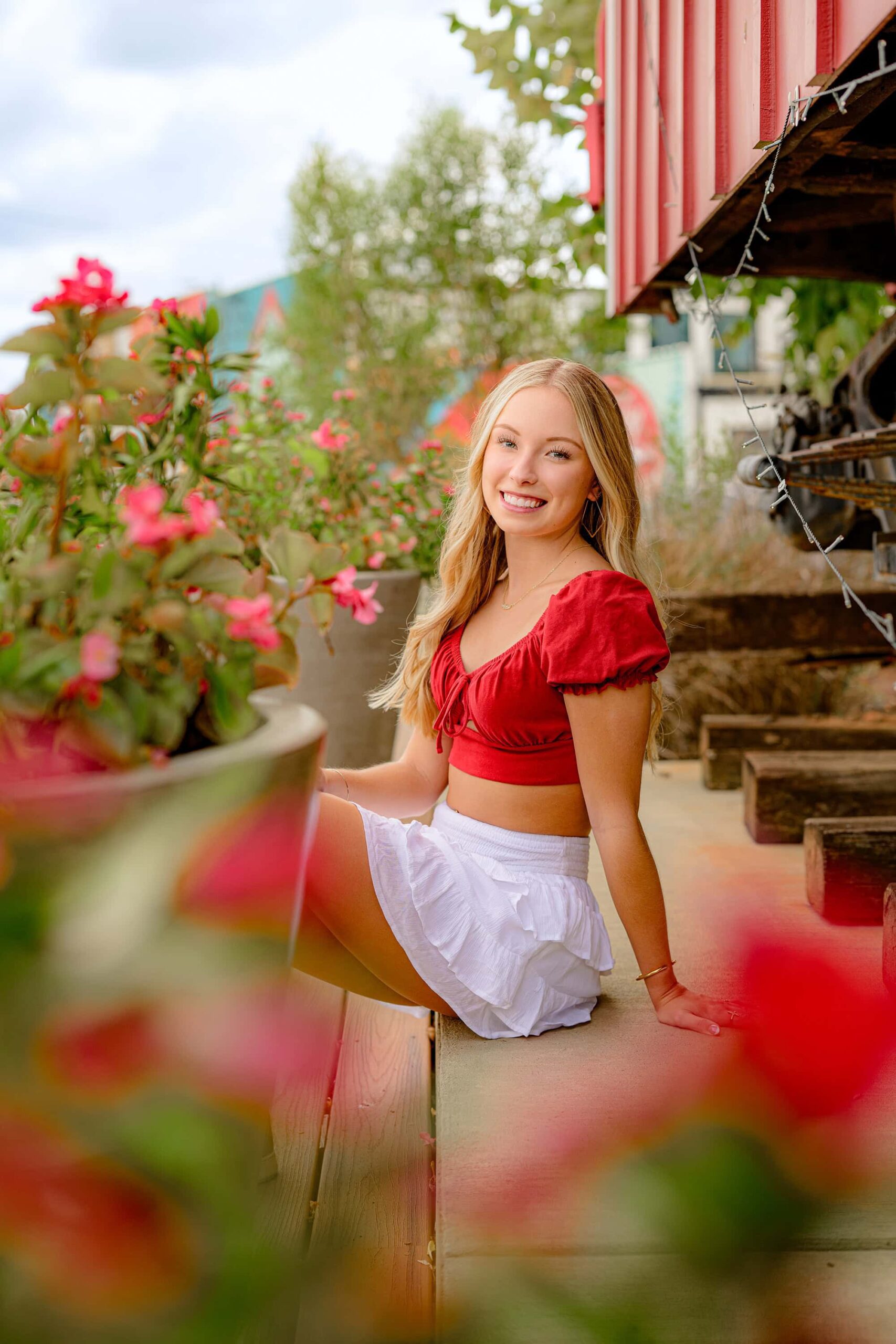 Waxhaw senior girl showcasing her style during an Essential Experience Grad Session, dressed in a white skirt, red crop top, and black-and-white Nike shoes, sitting confidently on a platform.