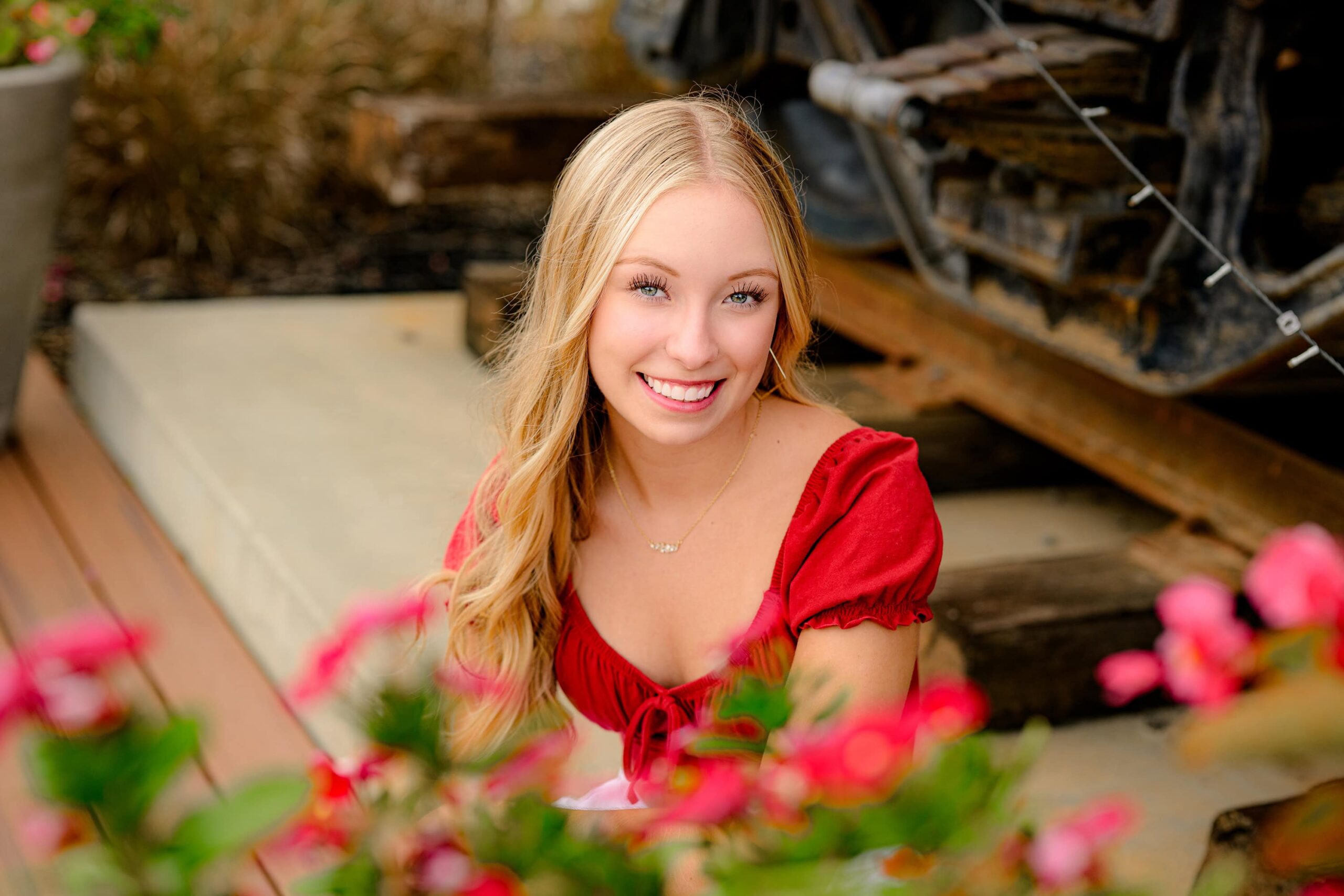 Charlotte, NC senior girl surrounded by vibrant flowers during an Essential Experience Grad Session, dressed in a white skirt and red crop top, sitting gracefully on a platform in Rock Hill, SC.
