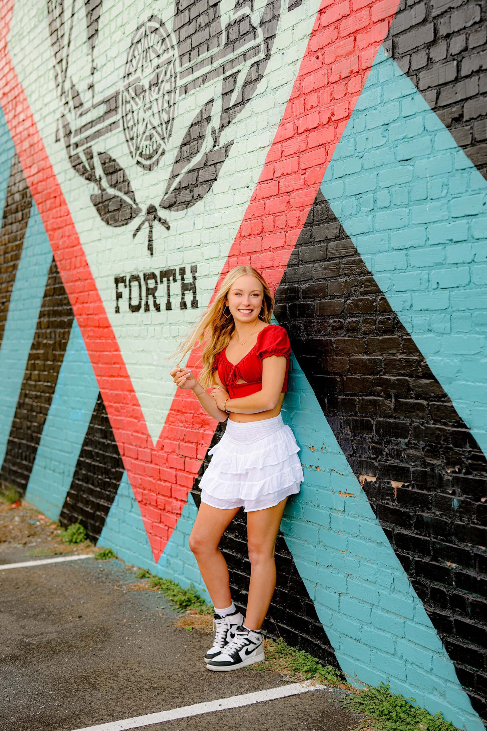Cuthbertson High School senior showcasing her personality during an Essential Experience Grad Session, dressed in a white skirt, red crop top, and black-and-white Nike shoes, standing confidently beside a vibrant graffiti wall.