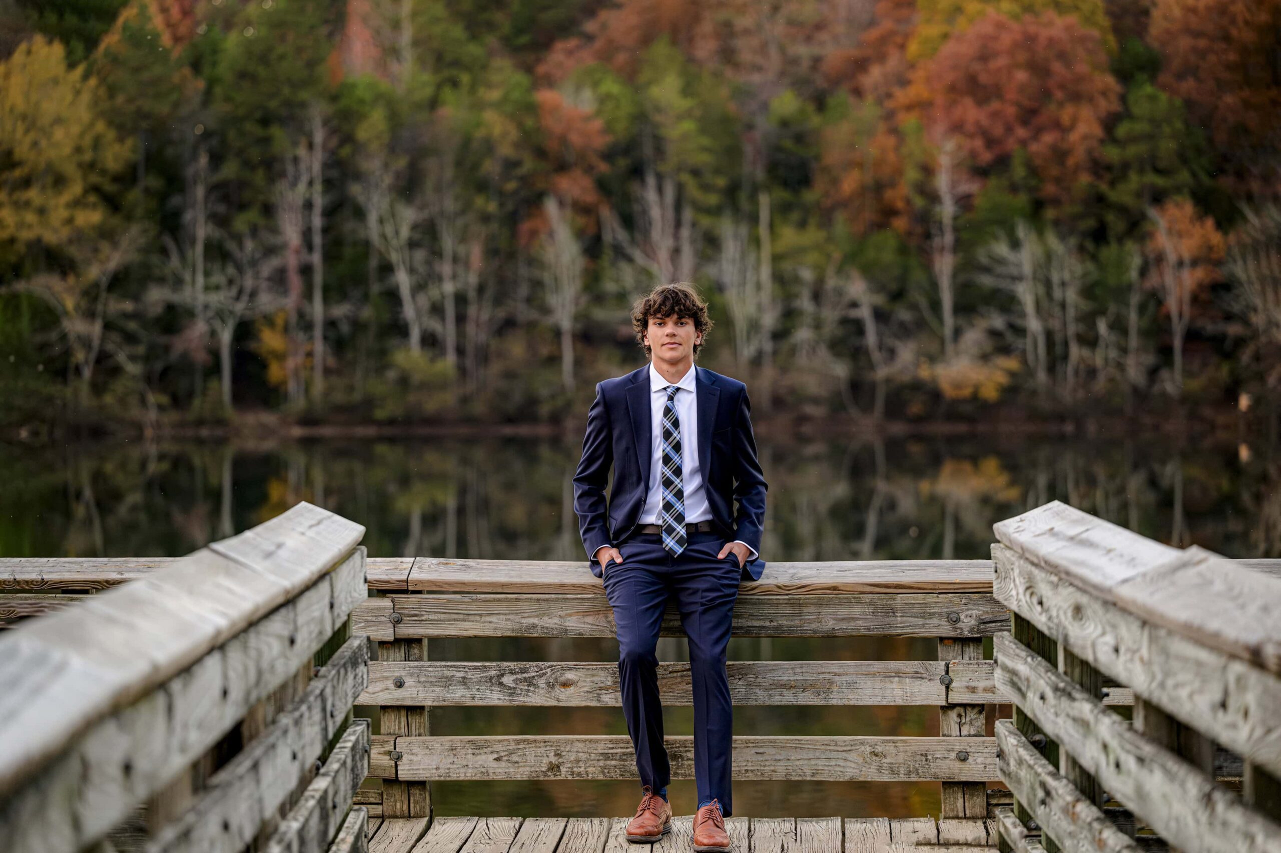boy sitting against a railing wearing a navy blue suit surrounded by fall color