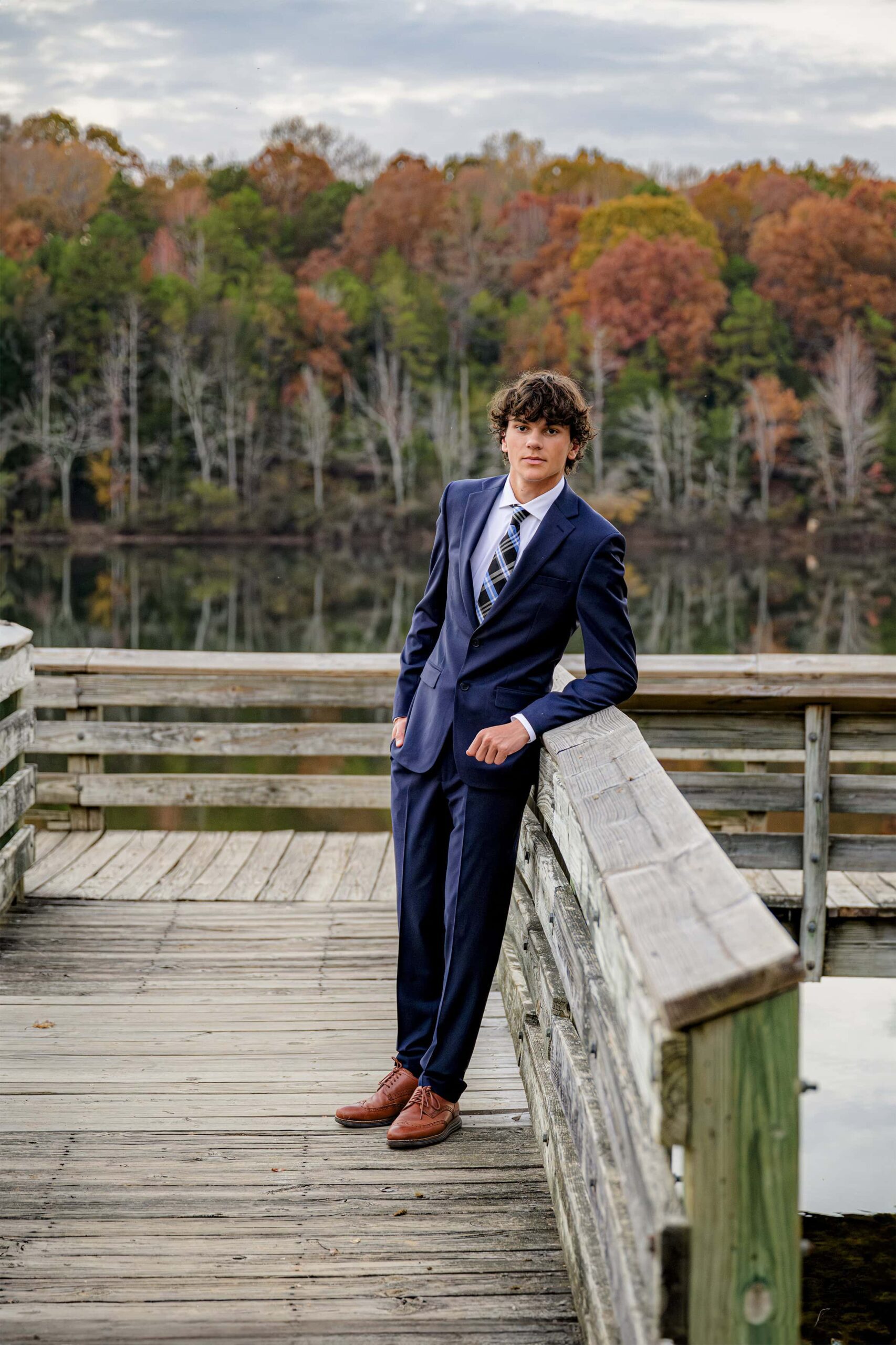 boy dressed in a dark navy suit with plaid tie and brown shoes leans against a railing with fall colors behind him