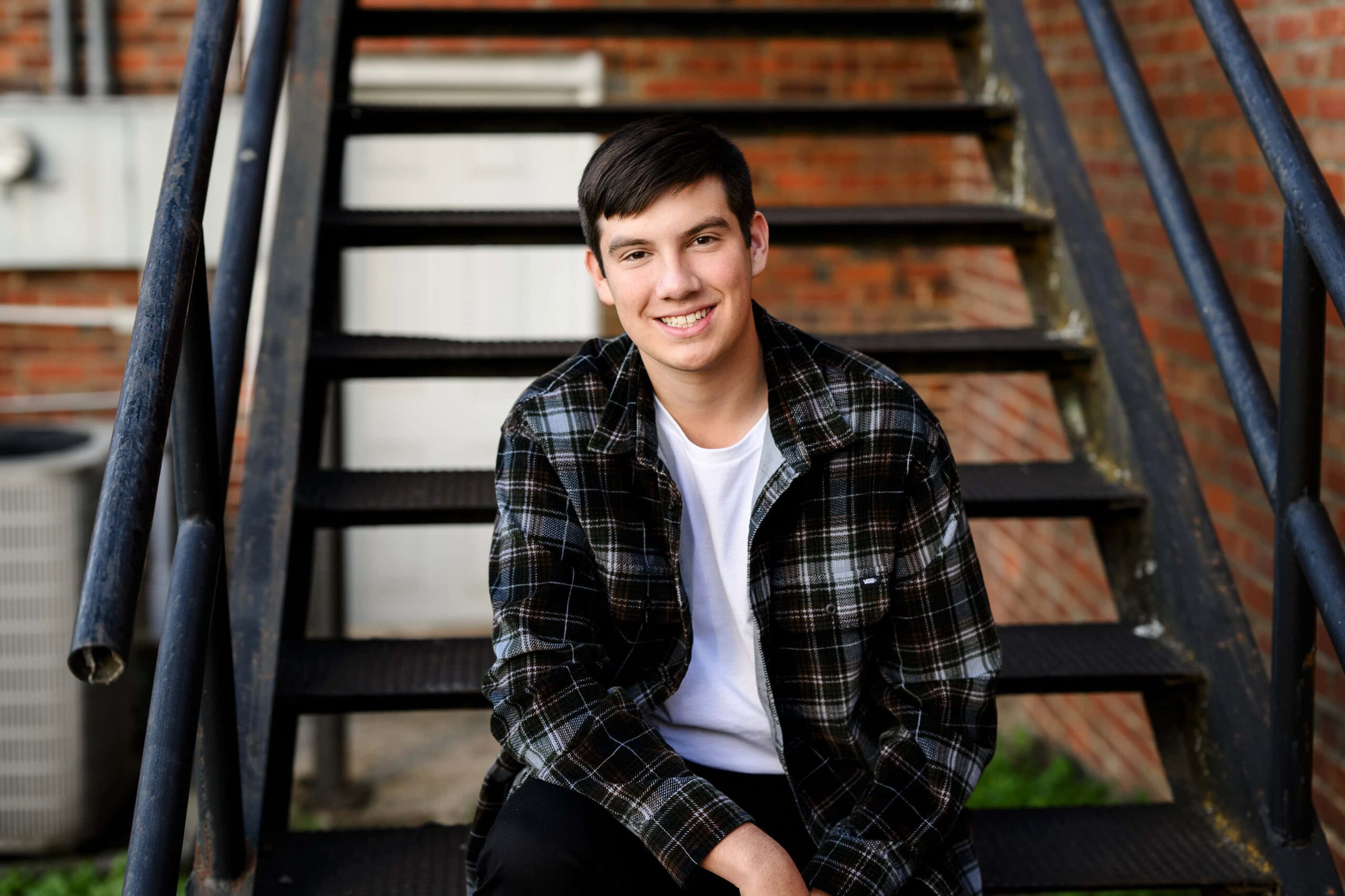 dark hair senior boy sitting on black metal staircase wearing a flannel shirt over a white t-shirt and jeans