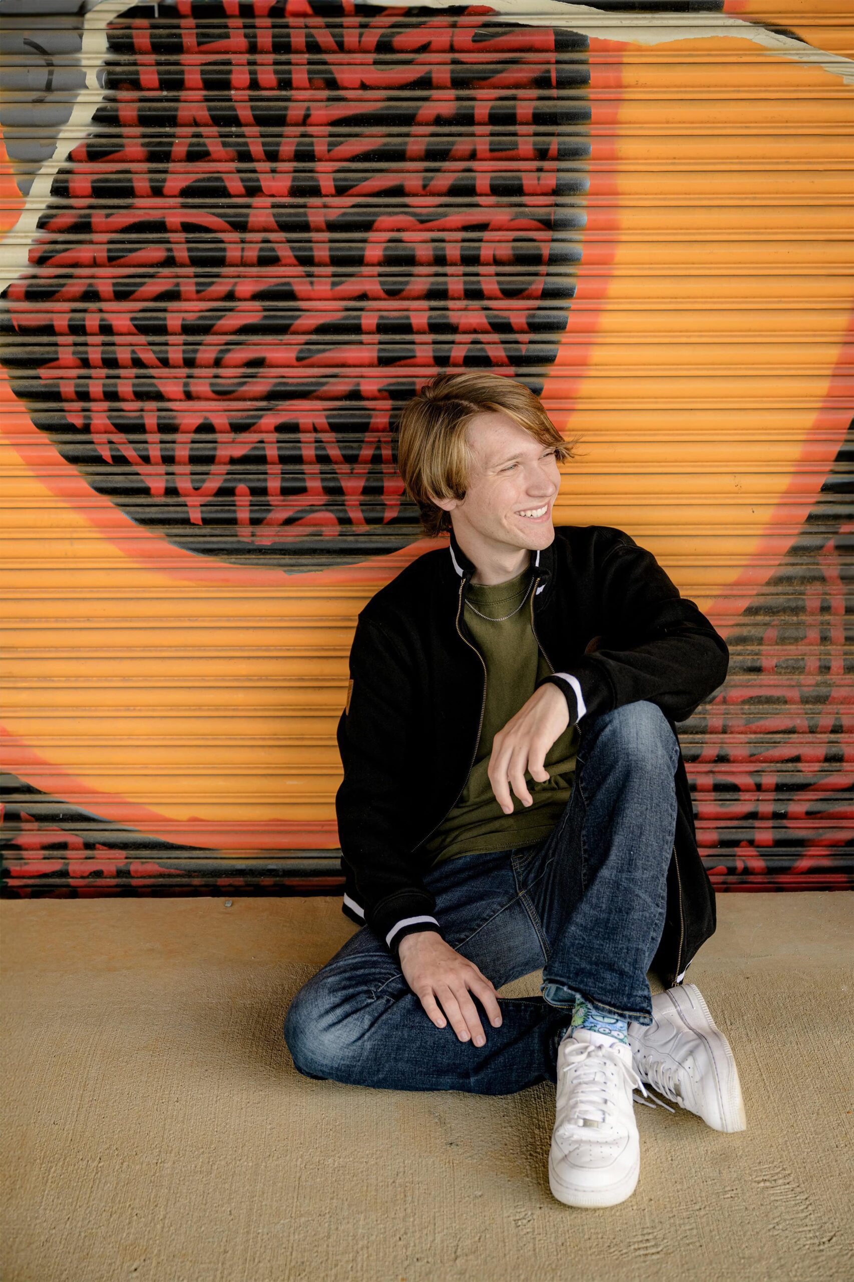 senior boy sits against a graphic wall wearing jeans a green shirt and tennis shoes