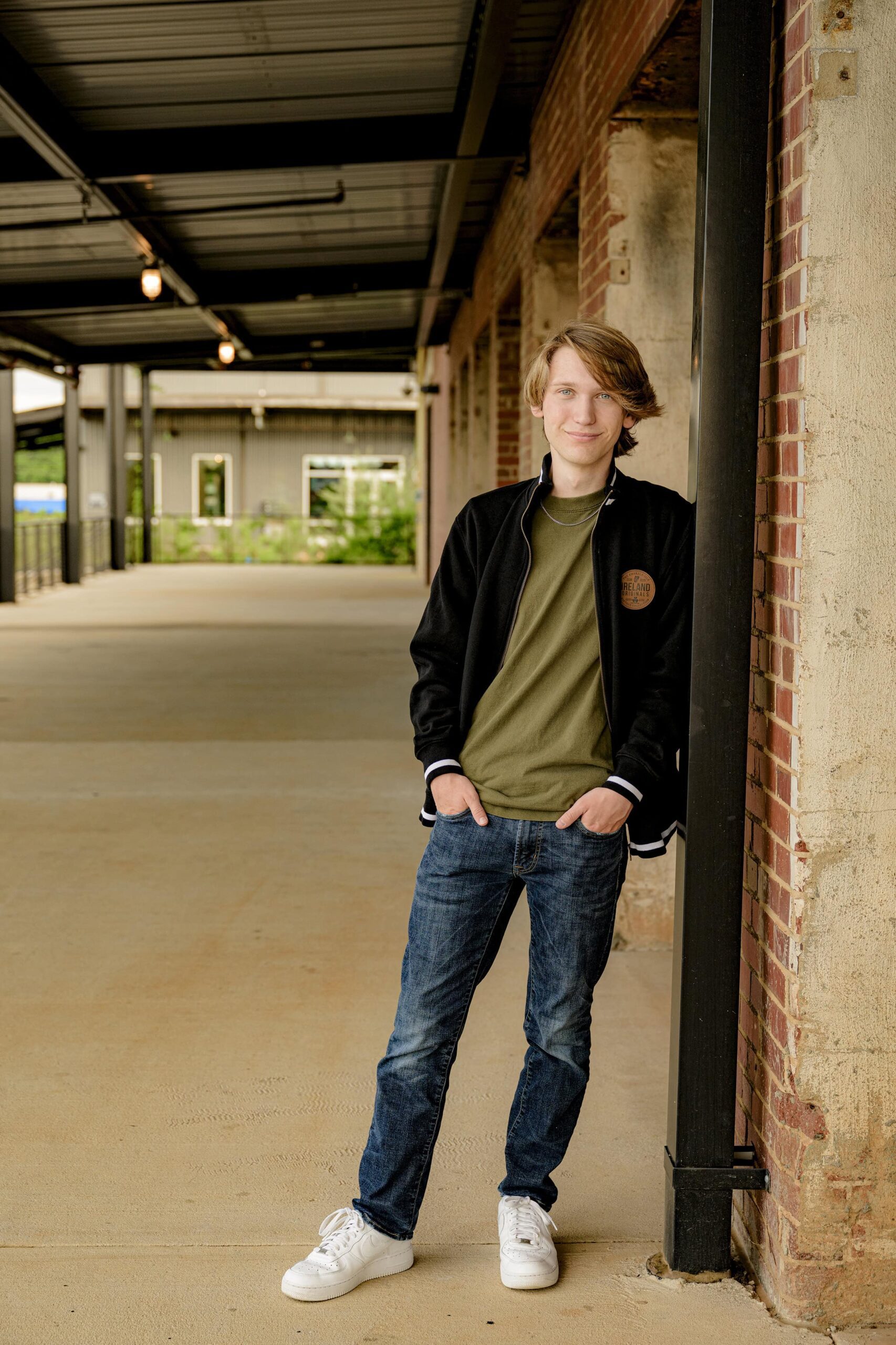 boy with styled longer hair wearing jeans, a t-shirt, shoes, and a black jacket leaning against a post in an urban setting