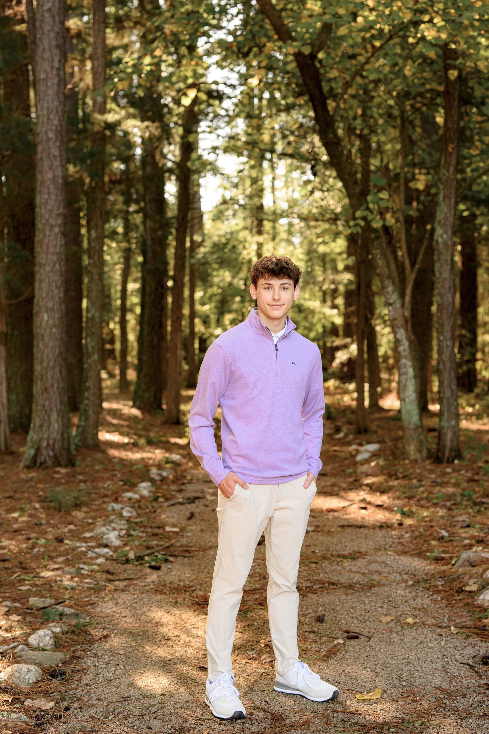 boy wears khaki pants and a lavender pullover standing in a tree lined pathway