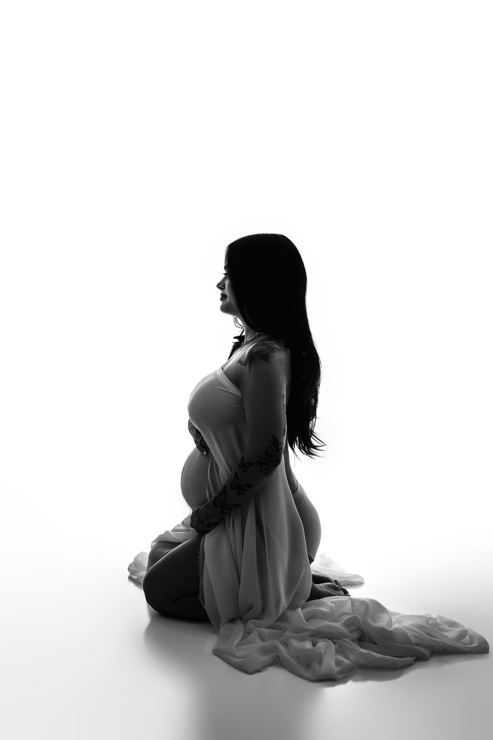 A beautifully intimate black and white portrait of the mama seated with sheer chiffon draped over her body. The fabric cascades around her, adding movement and a soft, elegant touch to this timeless maternity moment.