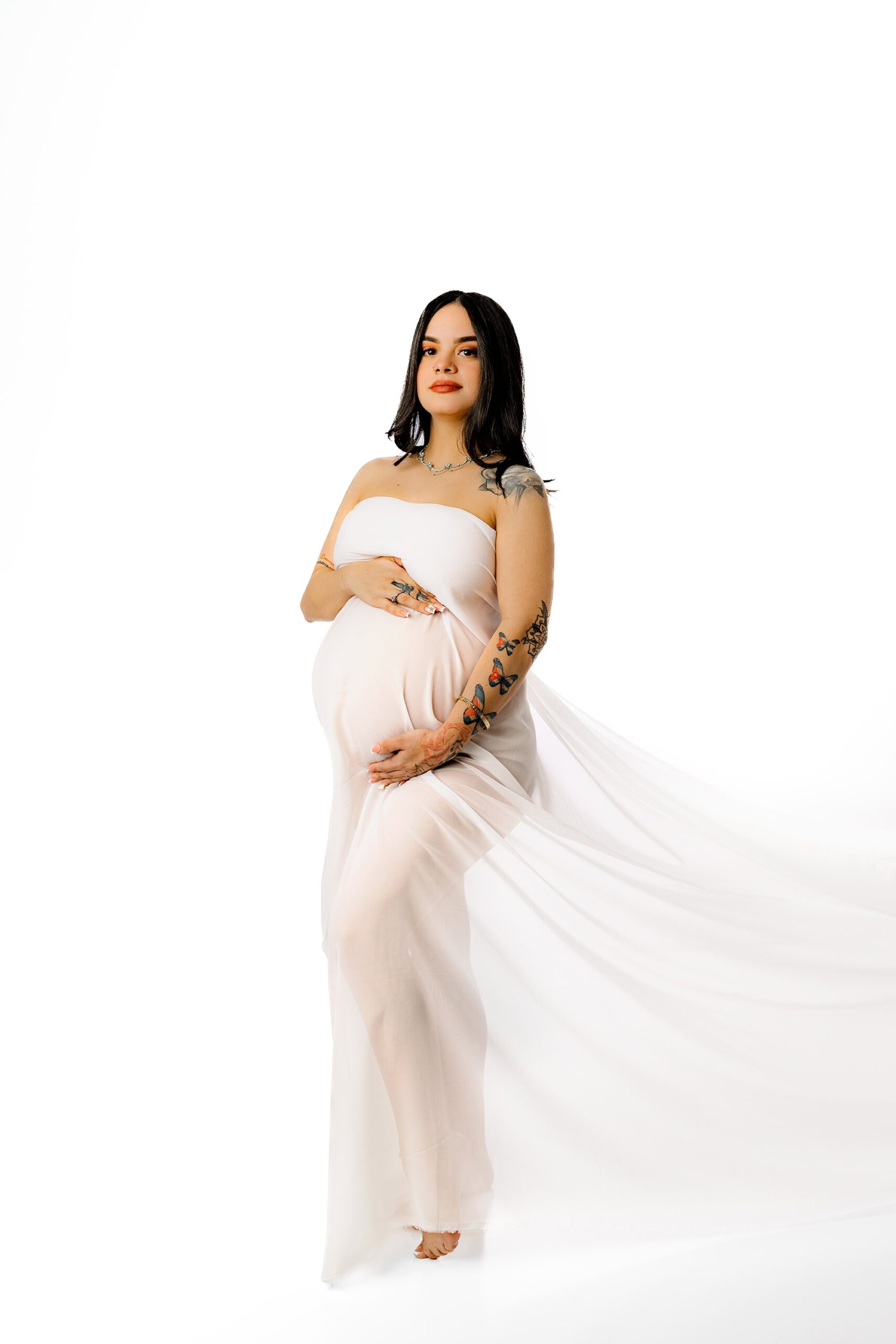 A breathtaking color image of the mama enveloped in sheer white chiffon, standing with her hands gently cradling her belly. The soft, flowing fabric and all-white studio setting create a dreamy, angelic aesthetic.