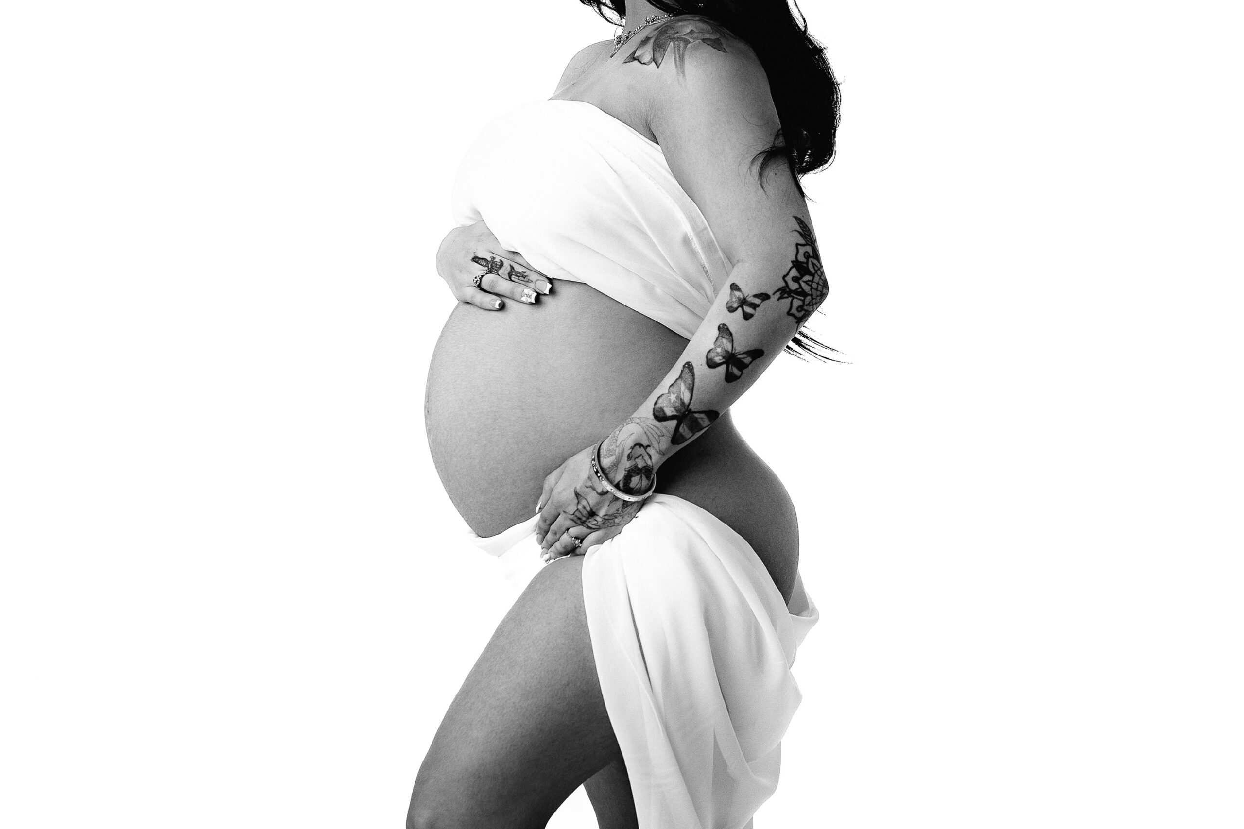 A striking black and white image capturing a dark-haired mama with tattoos, wrapped in soft white silk. The focus is on her belly and arms, emphasizing the delicate contrast between her inked skin and the flowing fabric.