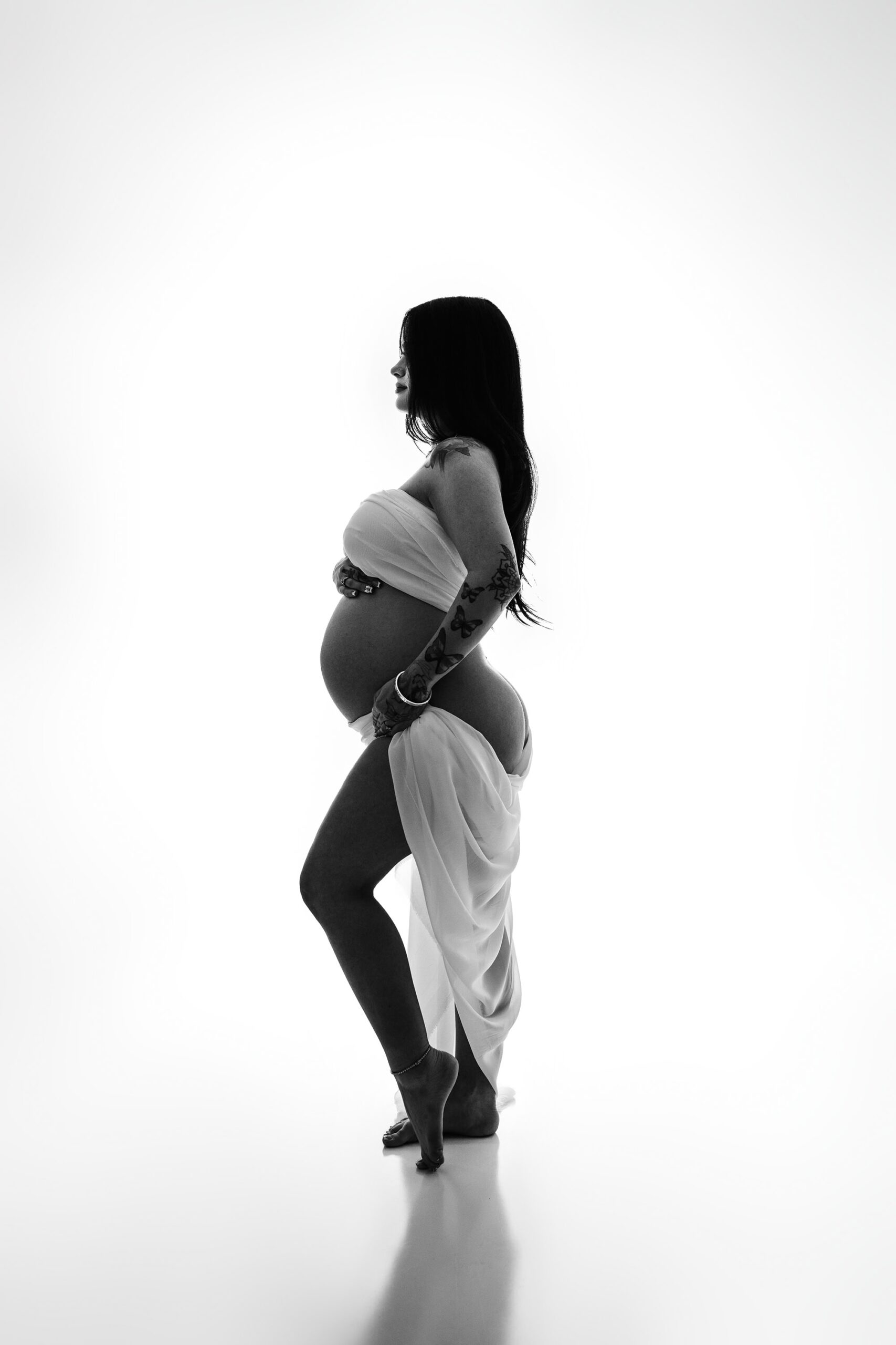 A graceful side-profile shot of the same mama, wrapped in sheer white chiffon. The fabric drapes elegantly around her breasts and under her backside, creating a timeless and artistic maternity portrait.