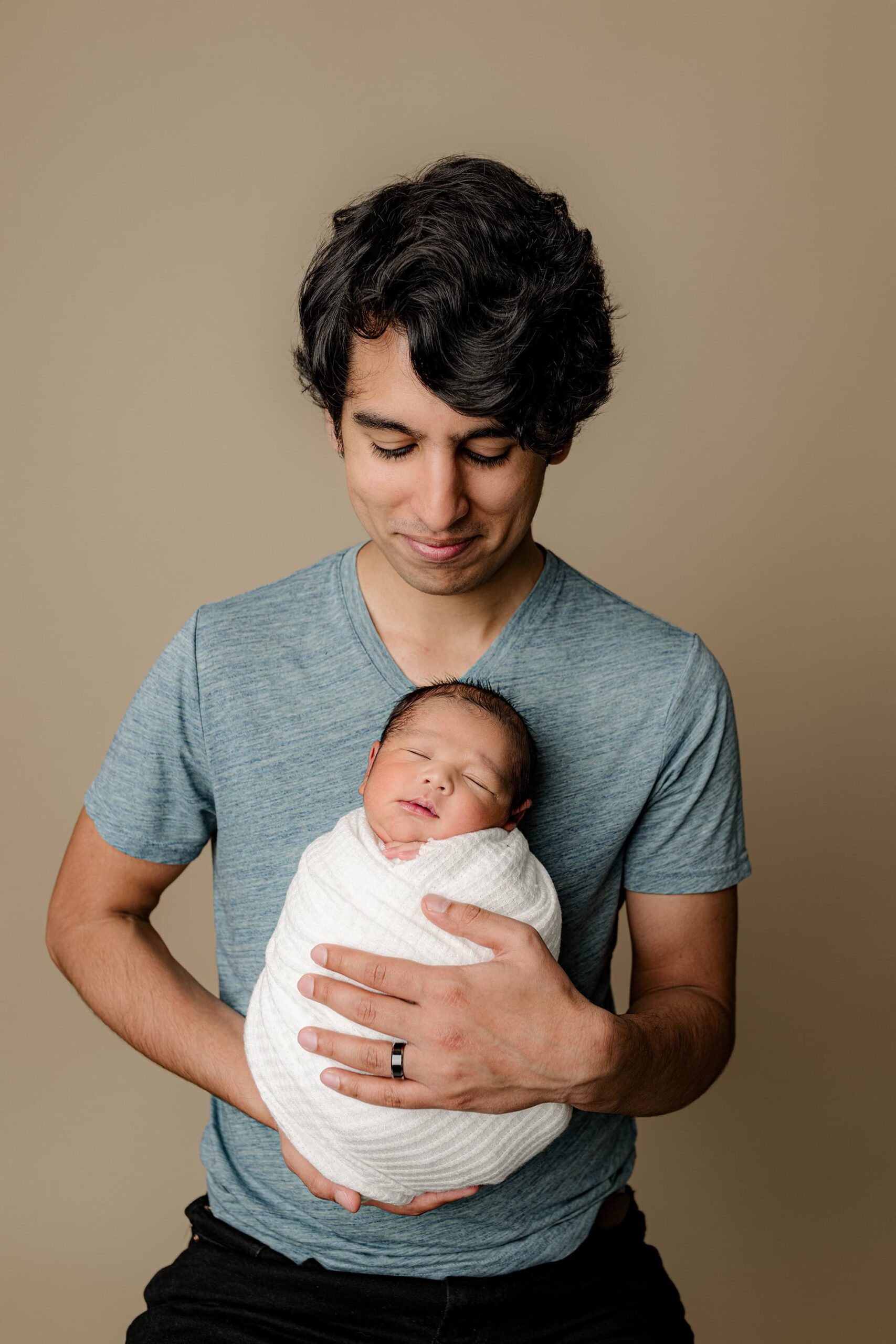 Charlotte area dad wears a blue shirt and is looking down at his newborn son wrapped in white