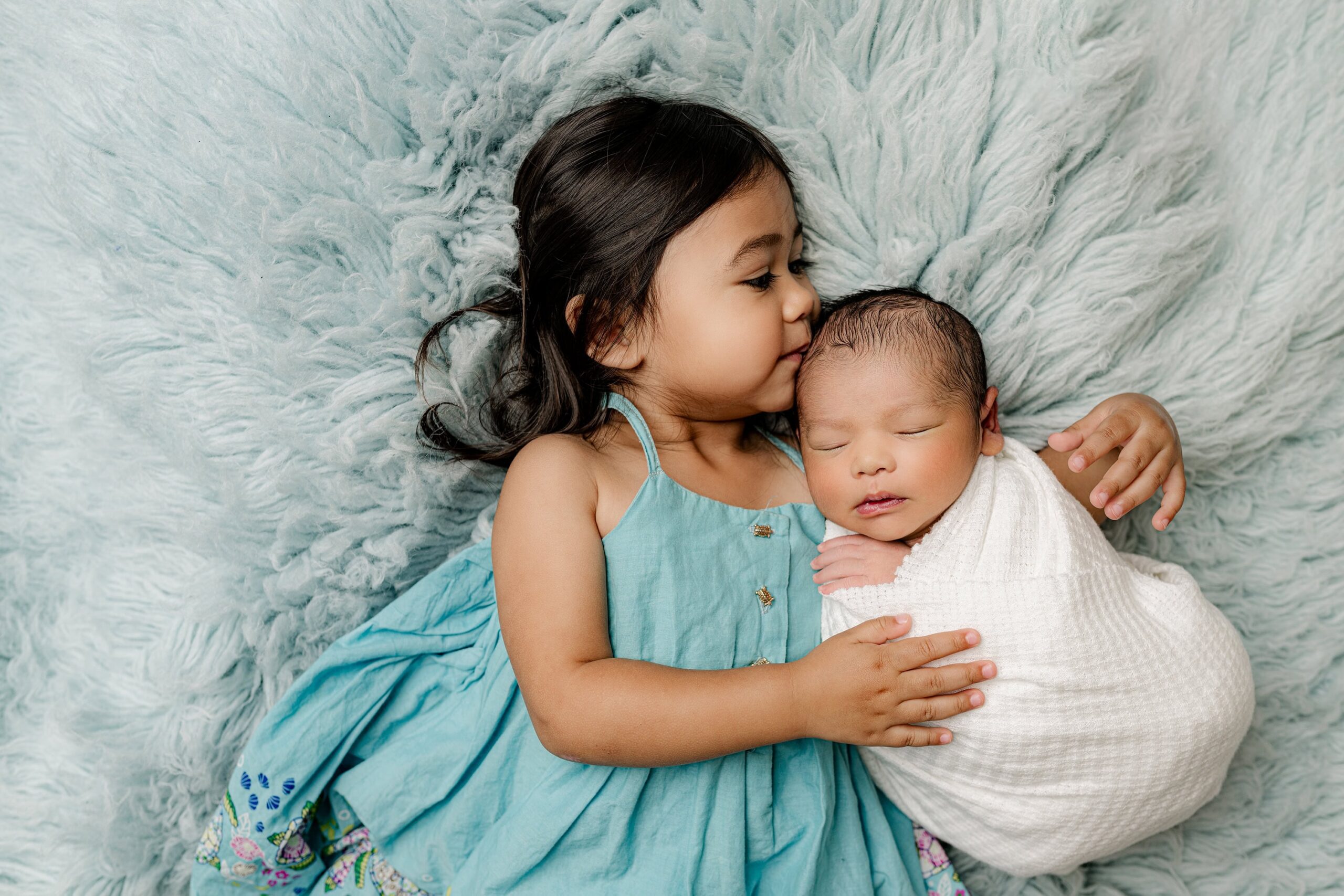 toddler sibling girl kisses the head of her newborn baby brother while laying on a blue fur in a waxhaw nc family newborn session