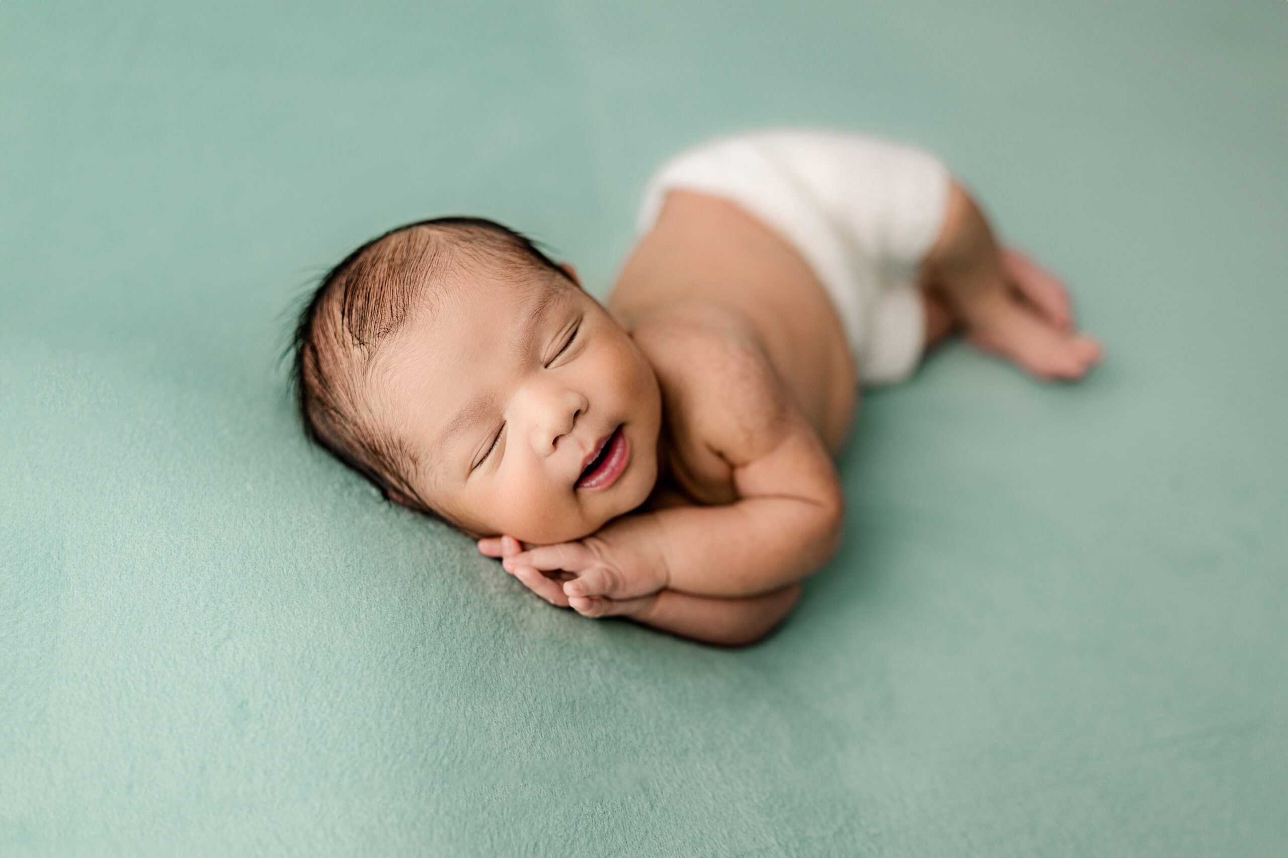 newborn baby boy with dark hair lays on his side with both hands under his cheek on a teal background during his family newborn session in a waxhaw, nc photography studio
