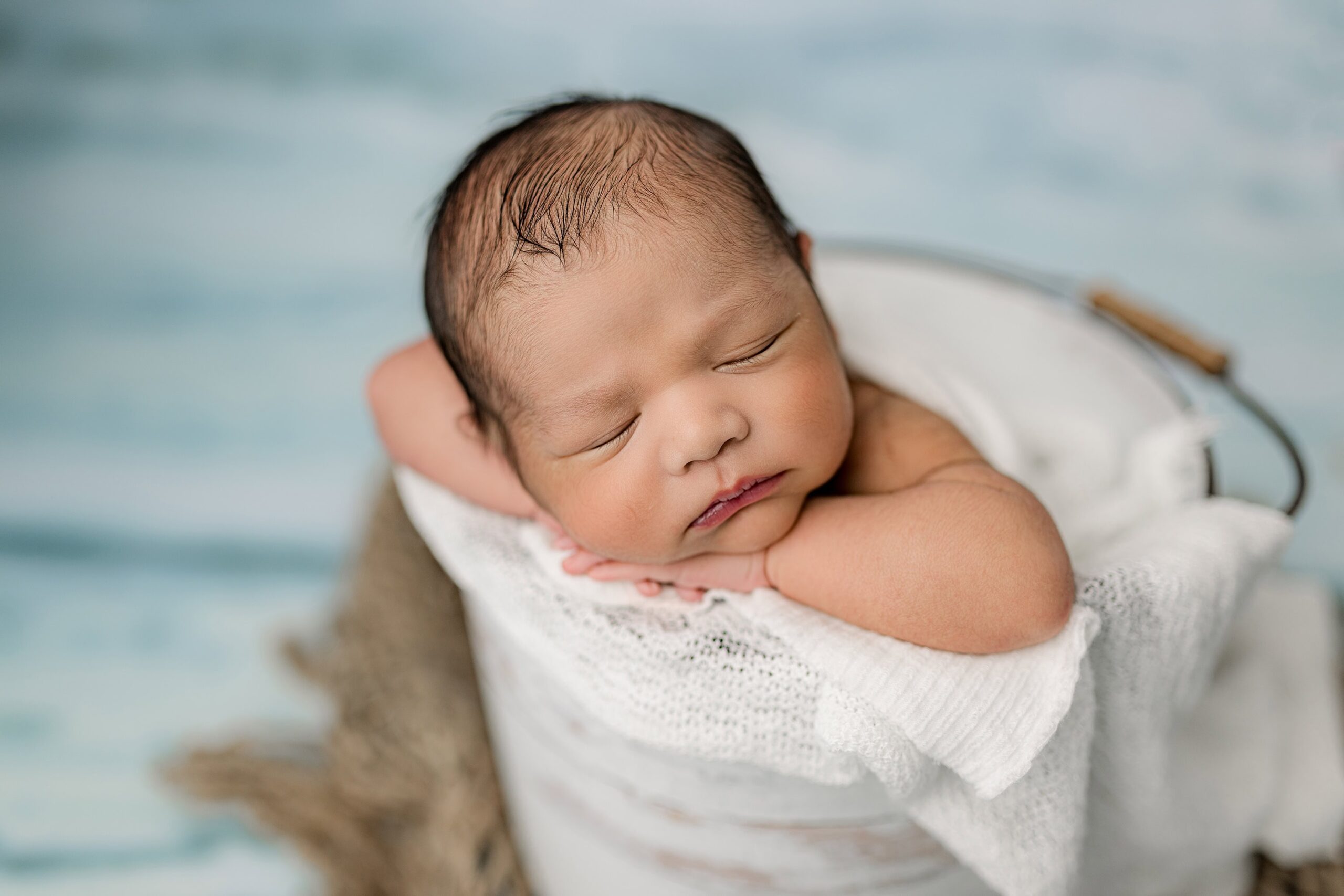 newborn boy is posed in a bucket with his hands under his chin on an ocean like background