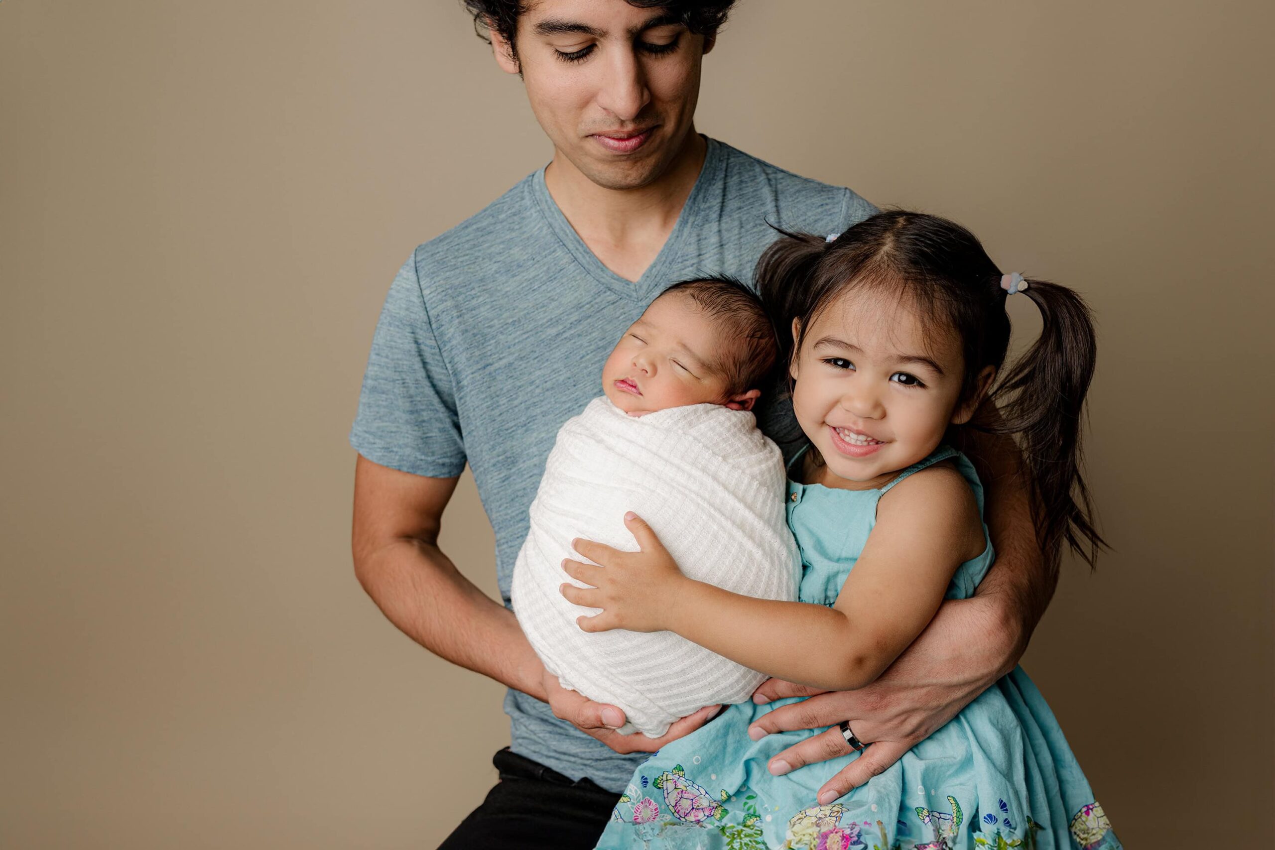dad is dressed in a blue shirt looking down as he holds newborn boy wrapped in white and his daughter dressed in blue dress