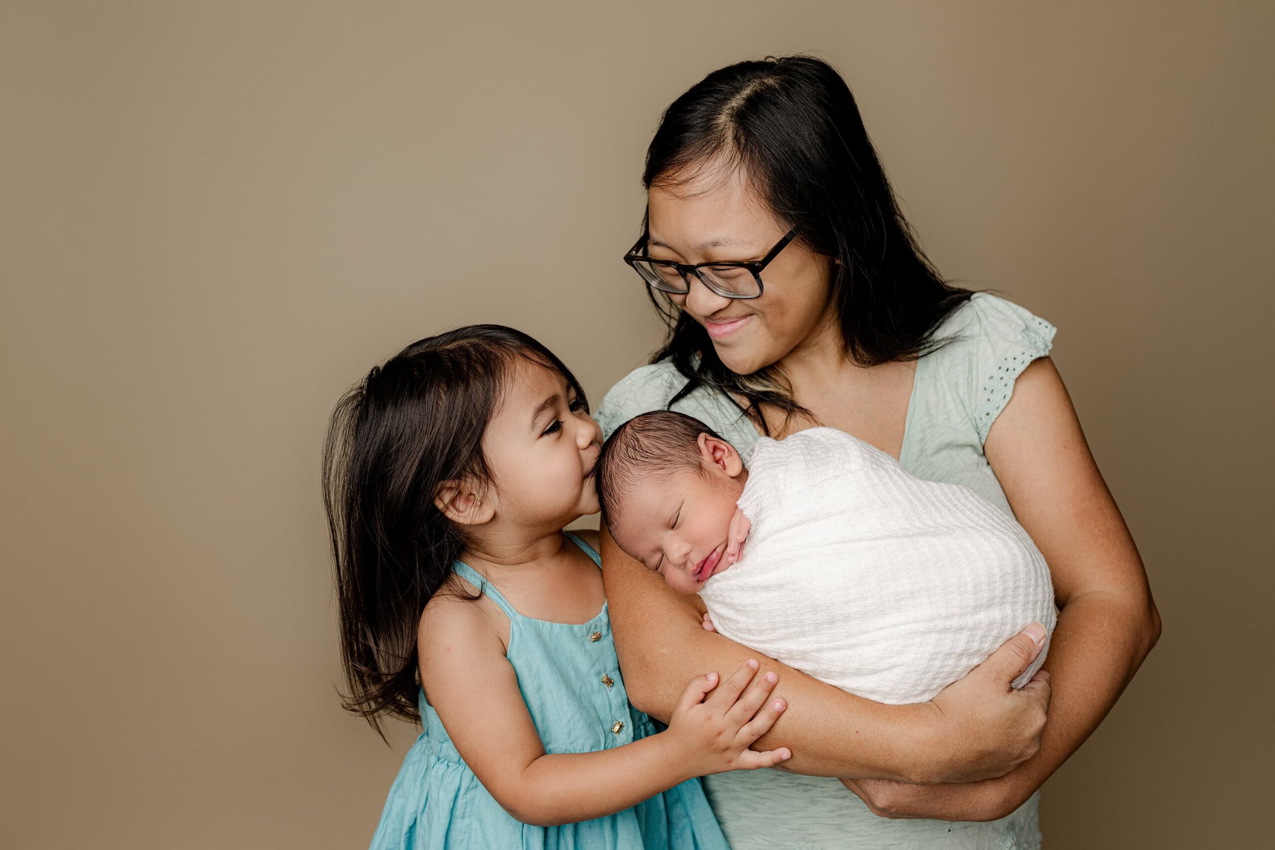 During their family newborn session big sister dressed in blue kisses the top of her little newborn brothers head while his mother is holding him