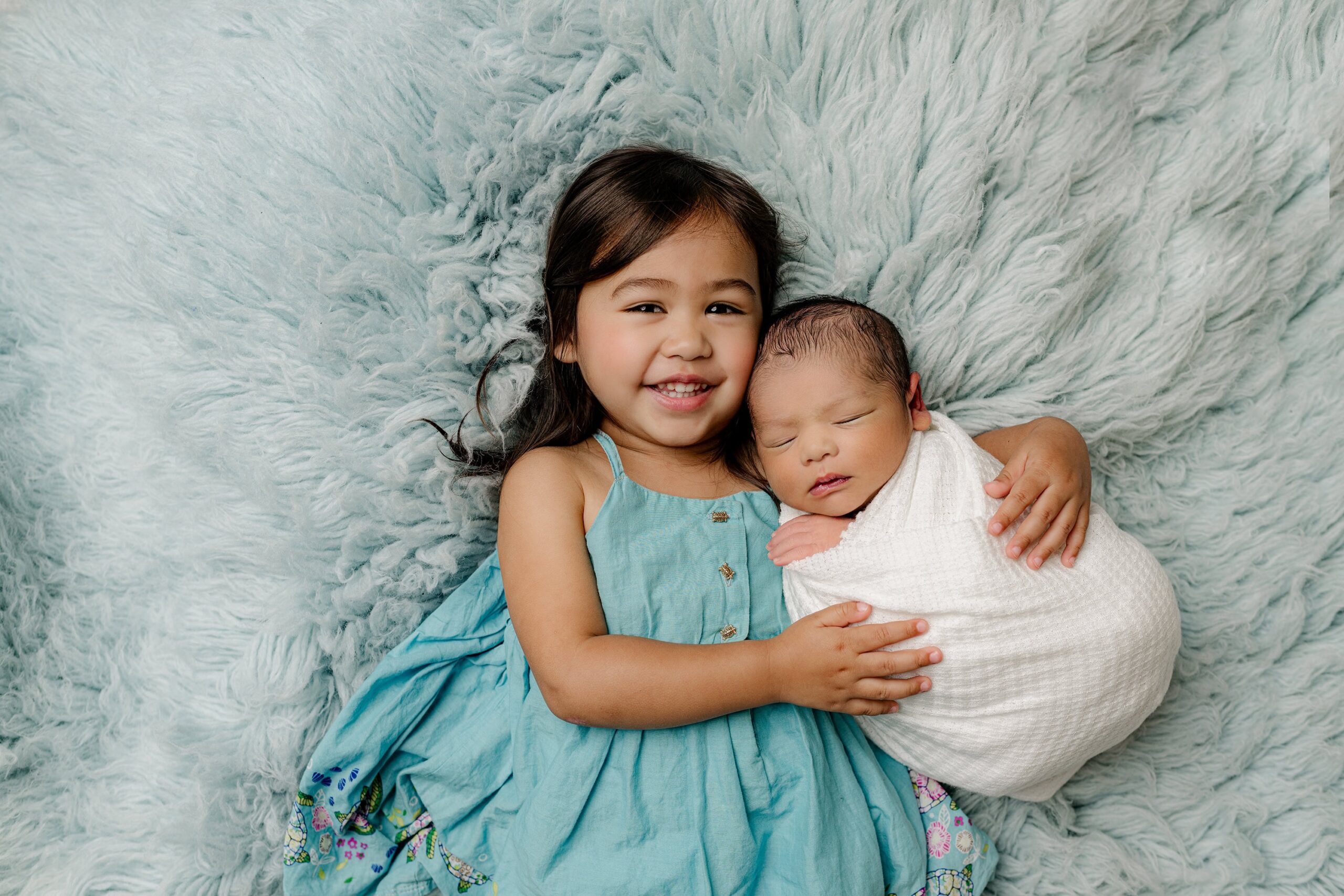 big sister dressed in shades of blue lays down holding her newborn brother wrapped in white on a blue rug in a waxhaw photo studio