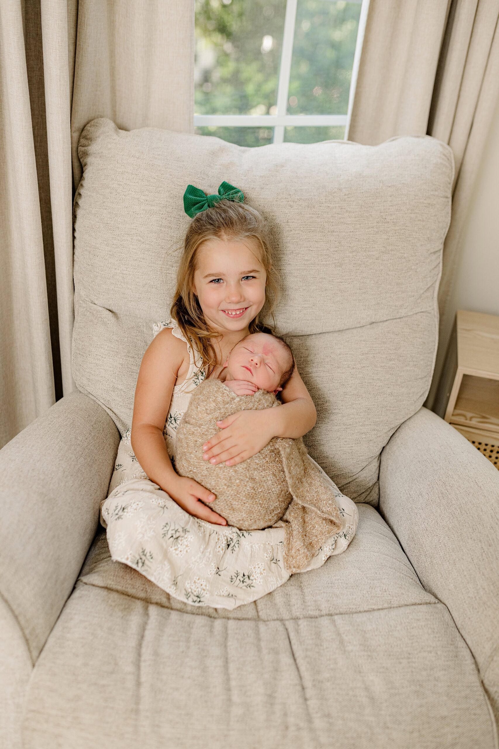 Sitting in a chair in the newborn nursery big sister wears a tan dress and green bow holding her brother wrapped in a brown wrap
