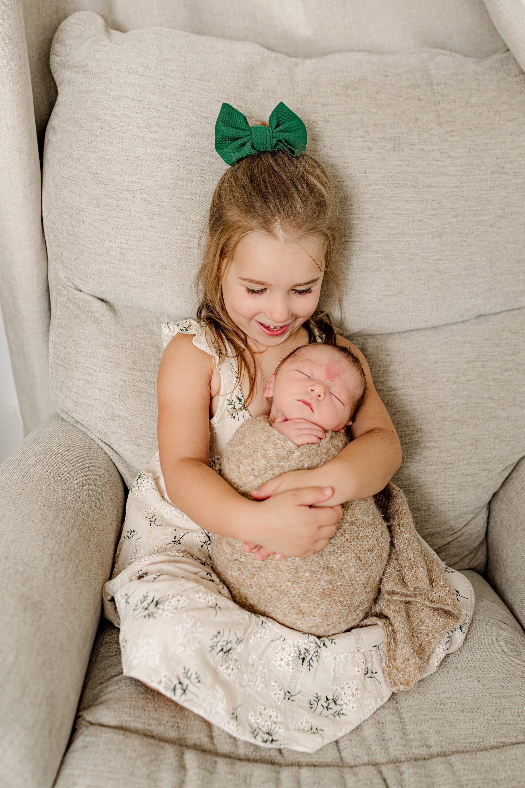 Big sister wears a tan dress and green bow in her blonde hair as she holds her newborn brother in her arms and looks down at him while sitting in a chair in his home nursery.