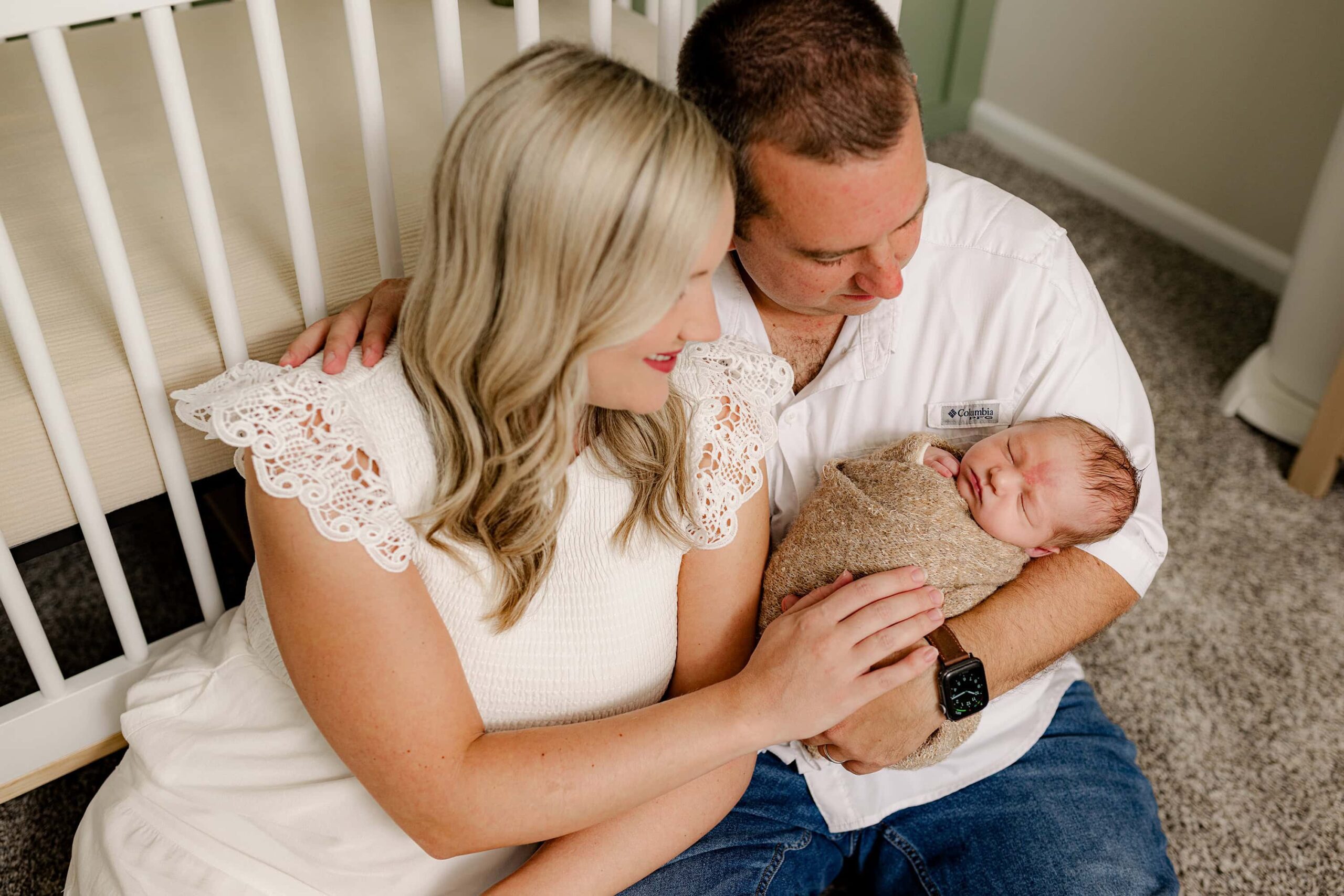 parents dressed in neutral tones look down at their newborn son wrapped in a brown knit wrap