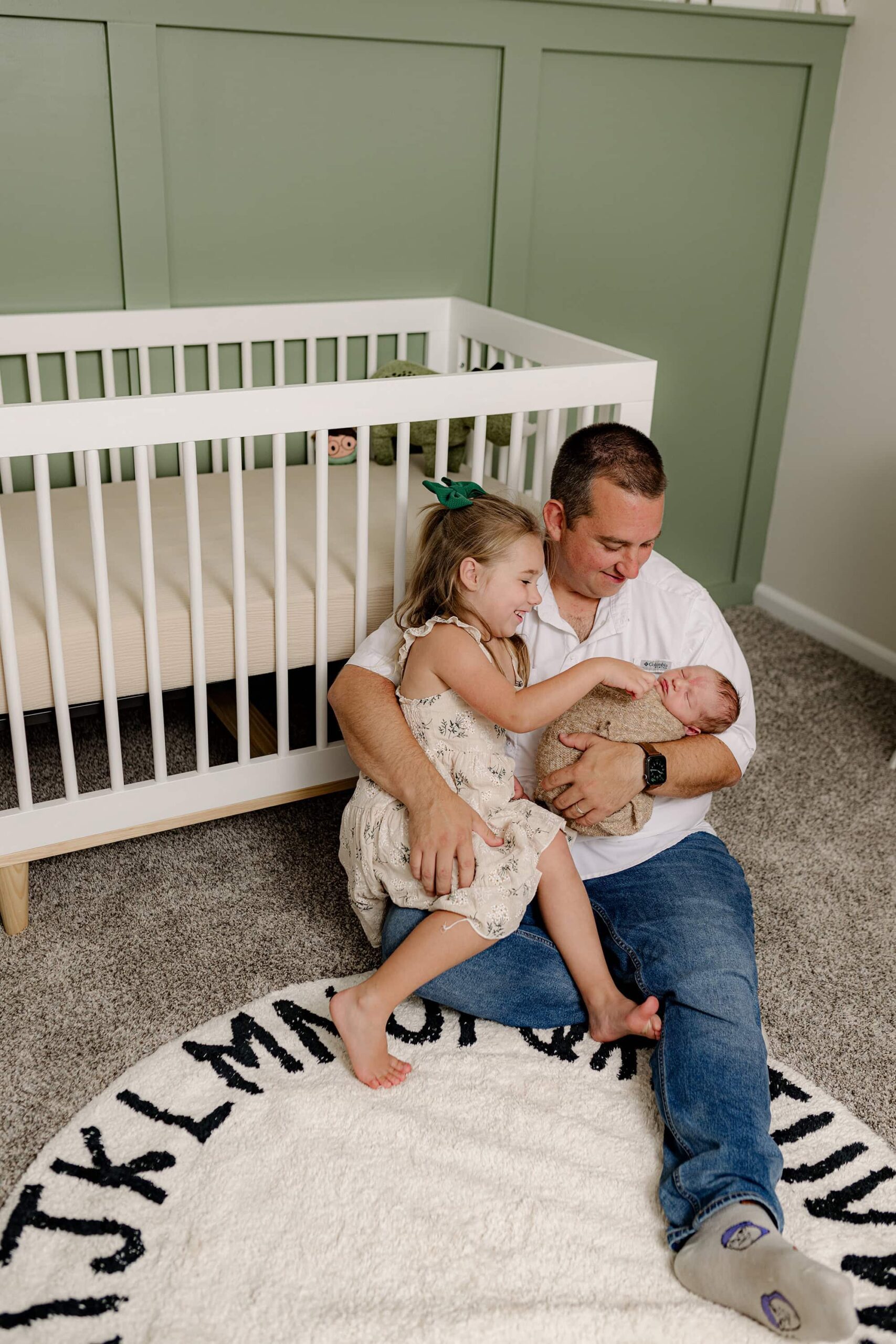new dad sits in the floor wearing jeans and a white shirt with holding his 5 year old daughter and newborn son in their home in indian land, sc
