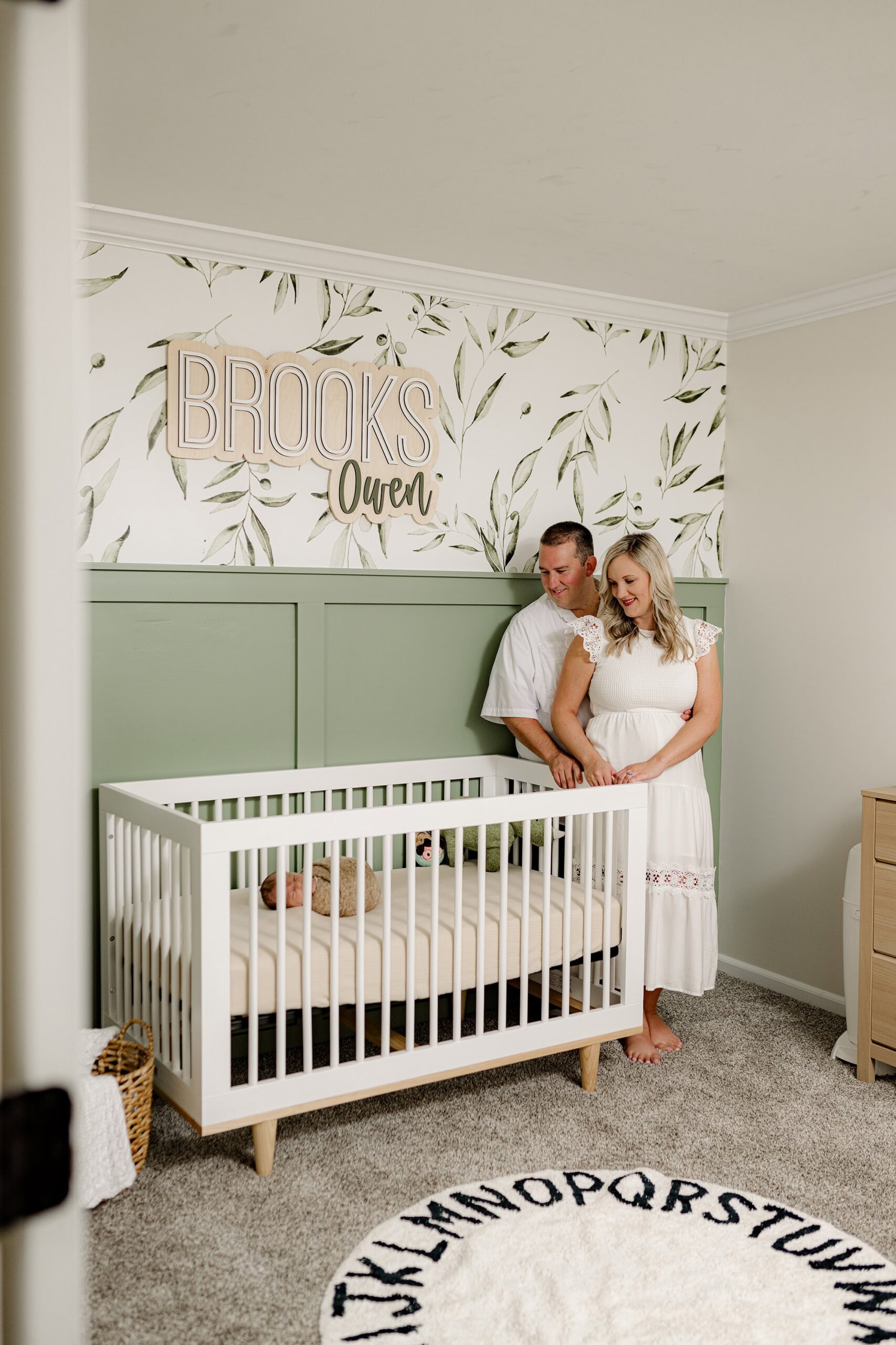 couple stands in their indian land, sc home nursery looking down at their son in his white crib in his naturally themed nursery with green walls, leaf based wall paper, and his name hung as a sign above his crib