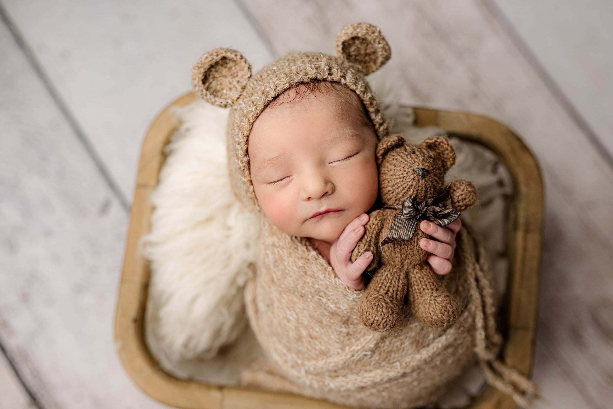 newborn boy is placed gently in a basket wrapped in a brown knit wrap with a matching bear bonnet and holding a brown teddy bear