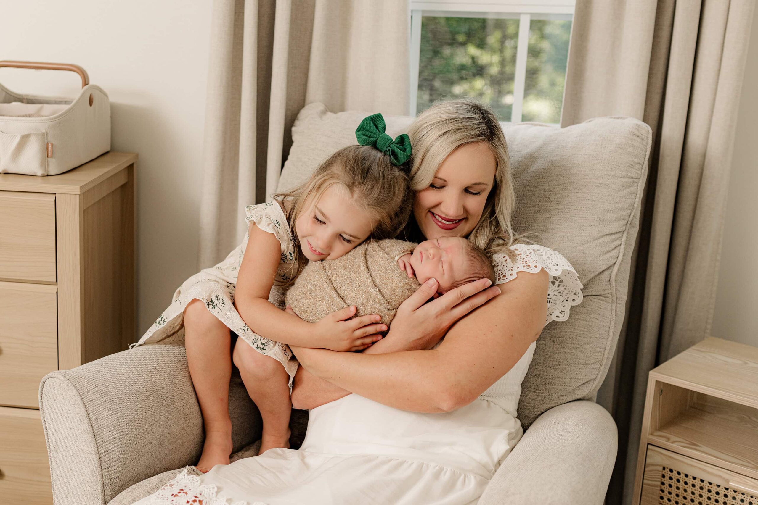 mother dressed in a cream dress sits in a chair smiling at her son as she also holds her daughter both cuddling the new baby