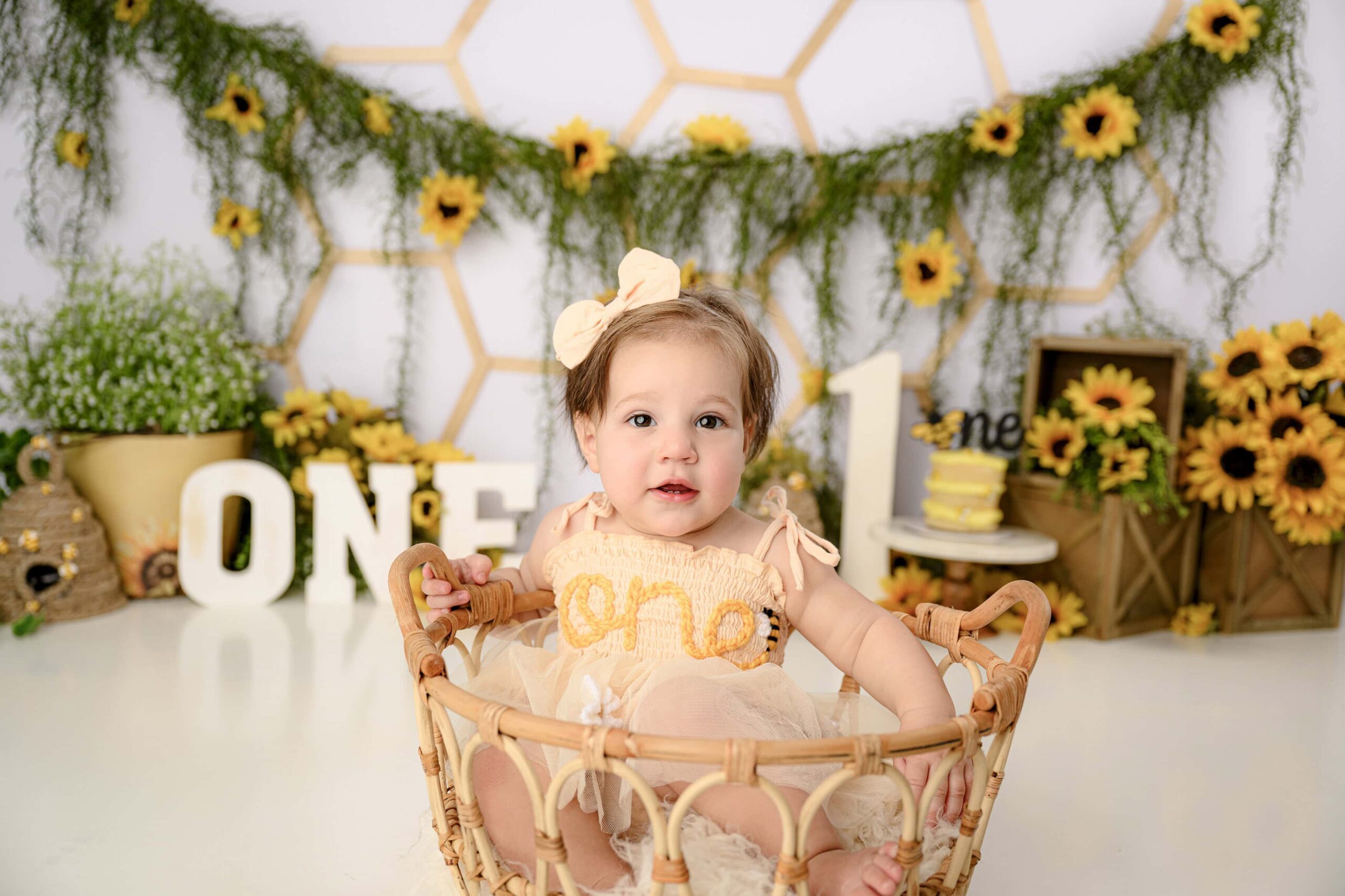 brown hair girl sitting in a basket during the start of her cake smash session dressed in a peach onsie and a coordinating peach bow