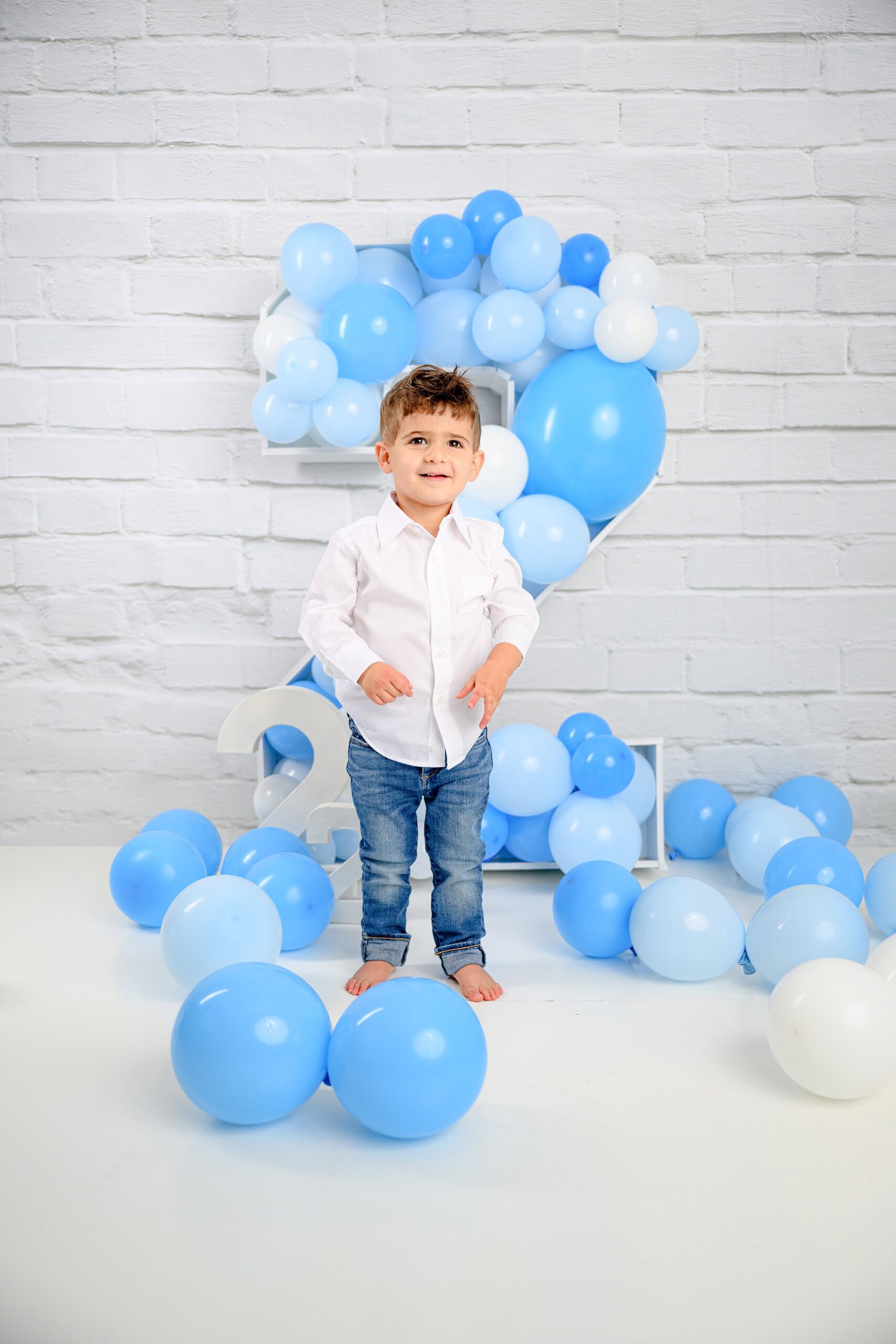 boy standing in his milestone session set surrounded balloons dressed in jeans and a white shirt