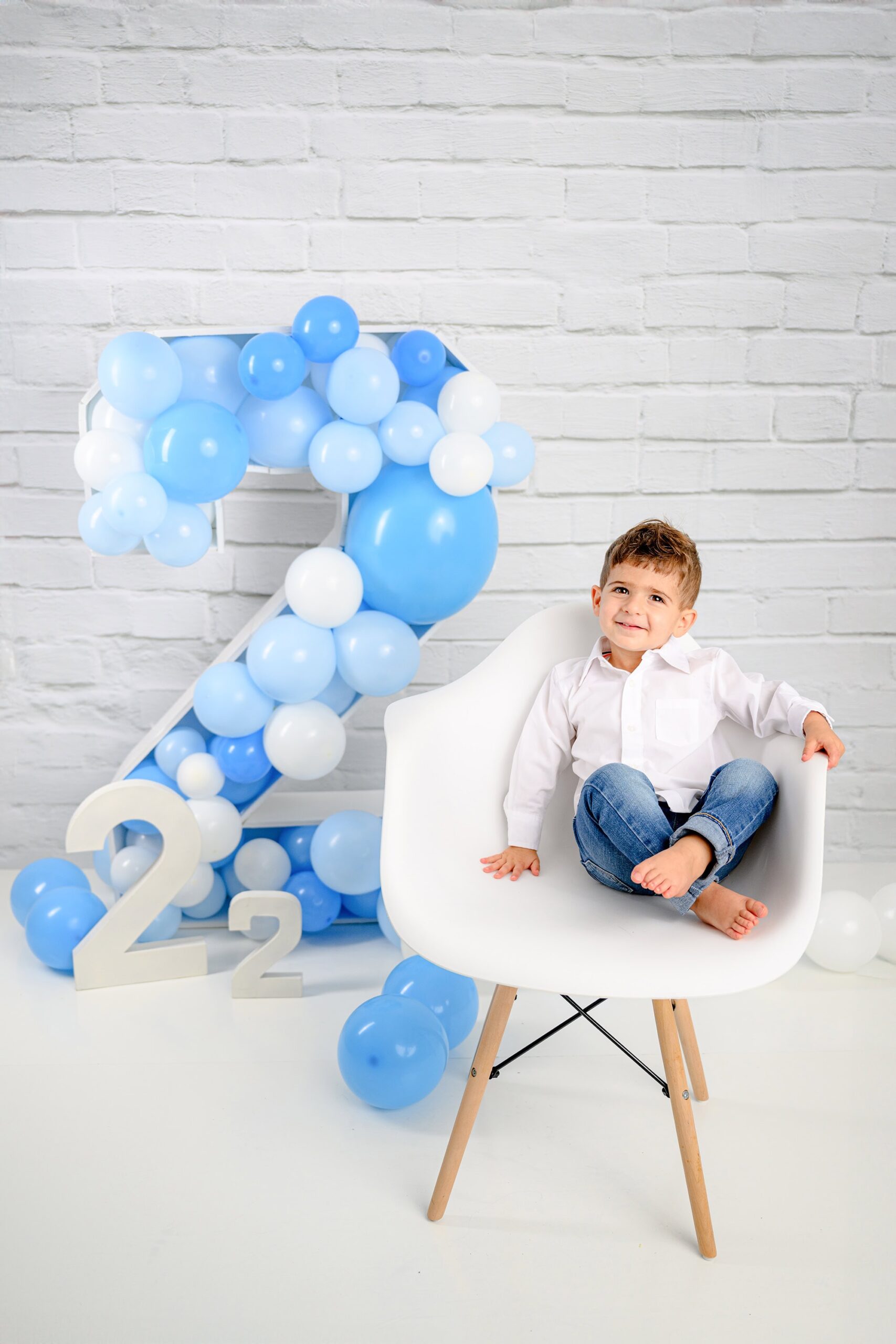 Boy wearing jeans and white shirt sitting in his chair during his two-year-old milestone session with a large number 2 prop filled with balloons
