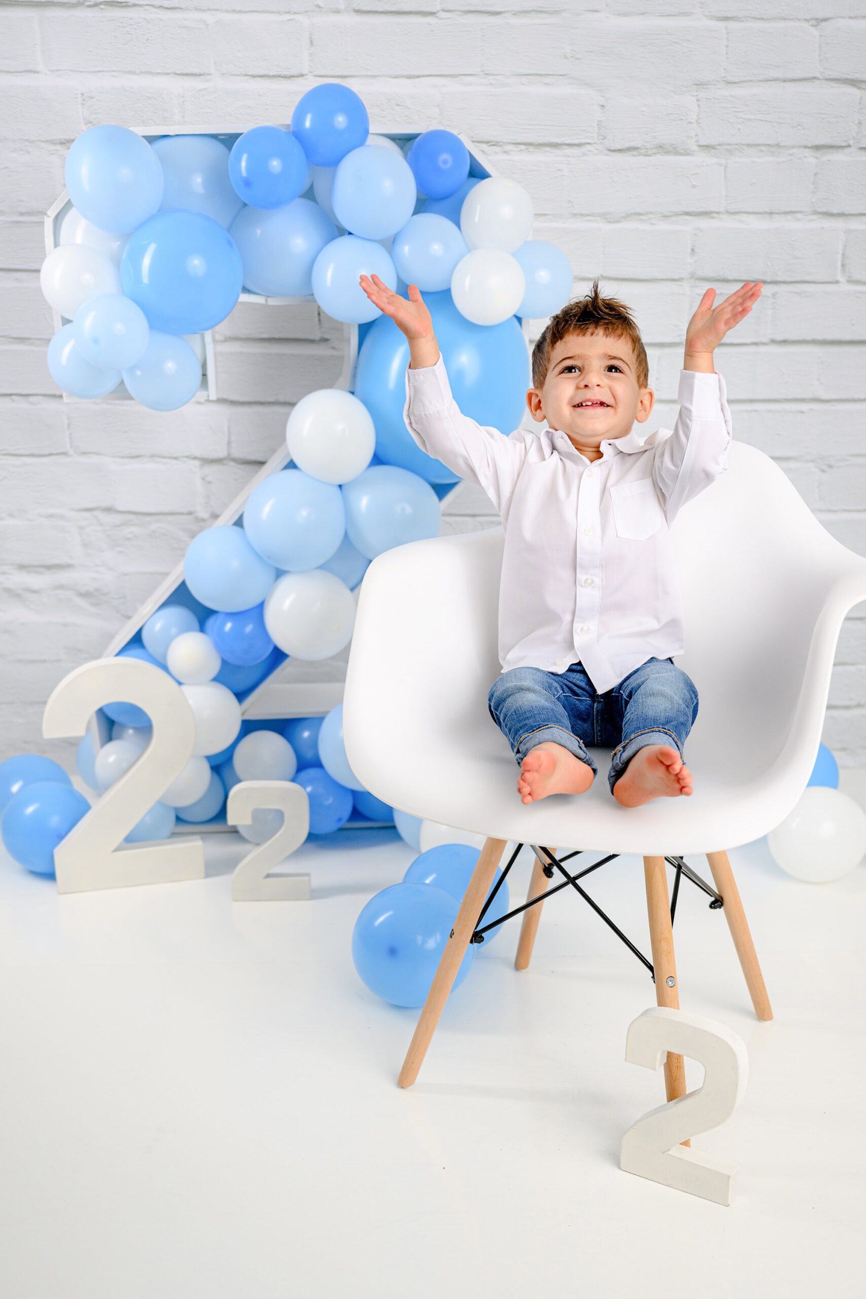 Wearing jeans and white shirt sitting in his chair during his two-year-old milestone session with a large number 2 prop filled with balloons