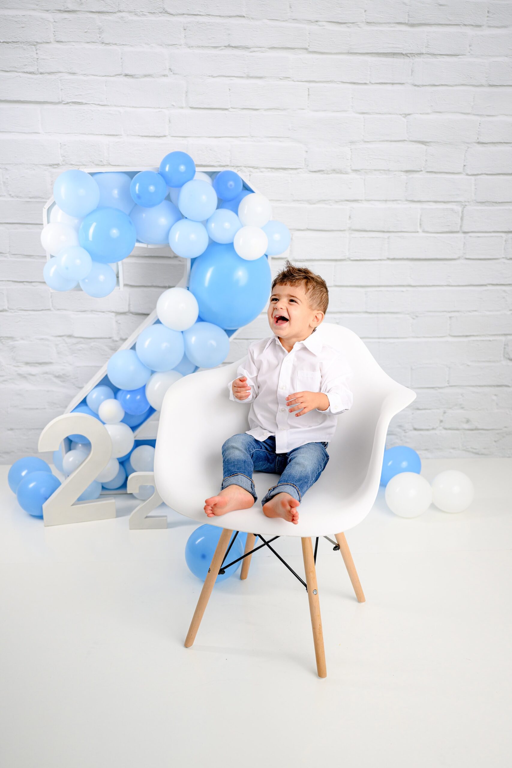 Wearing jeans and white shirt sitting in his chair during his two-year-old milestone session with a large number 2 prop filled with balloons