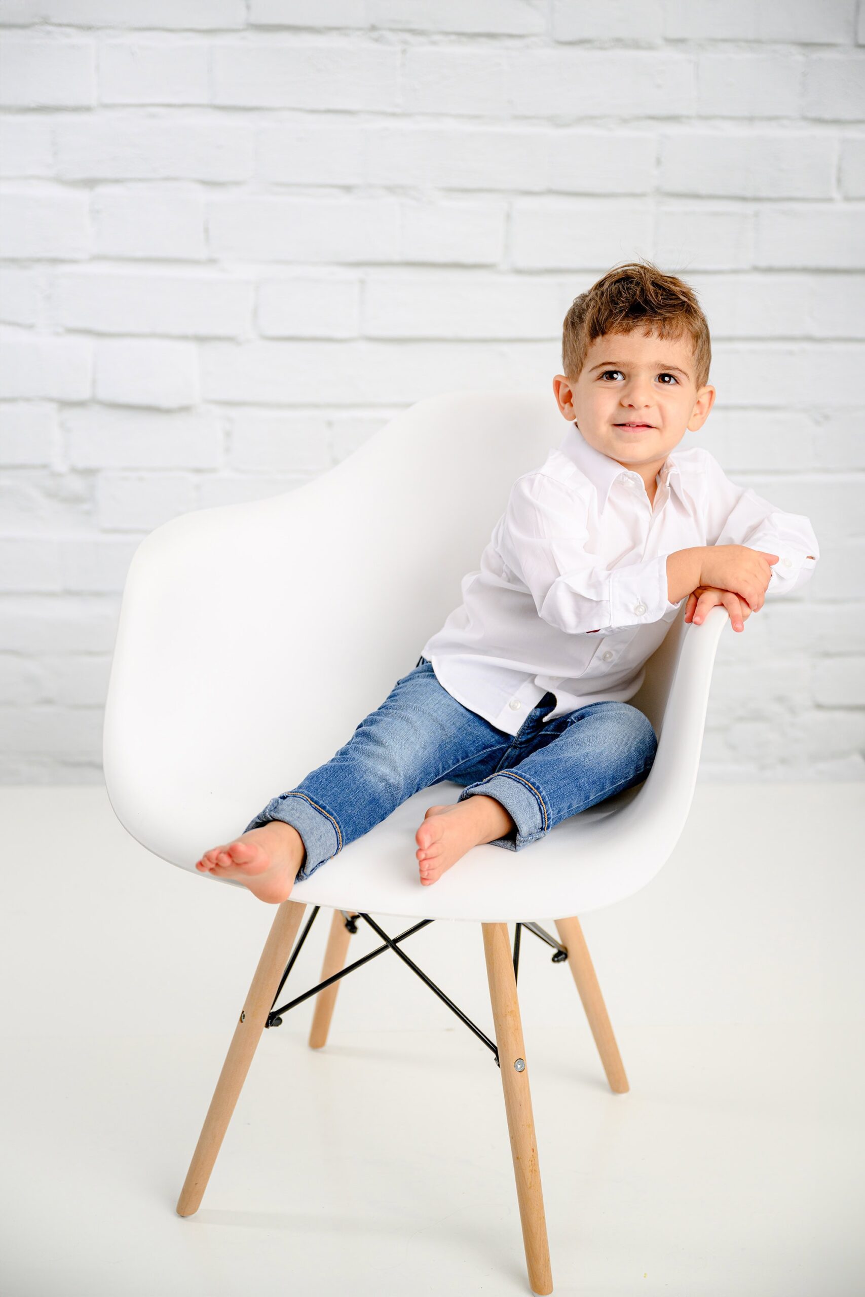 little boy sits in chair wearing a white shirt and jeans with a brick white background during his two year old milestone birthday session