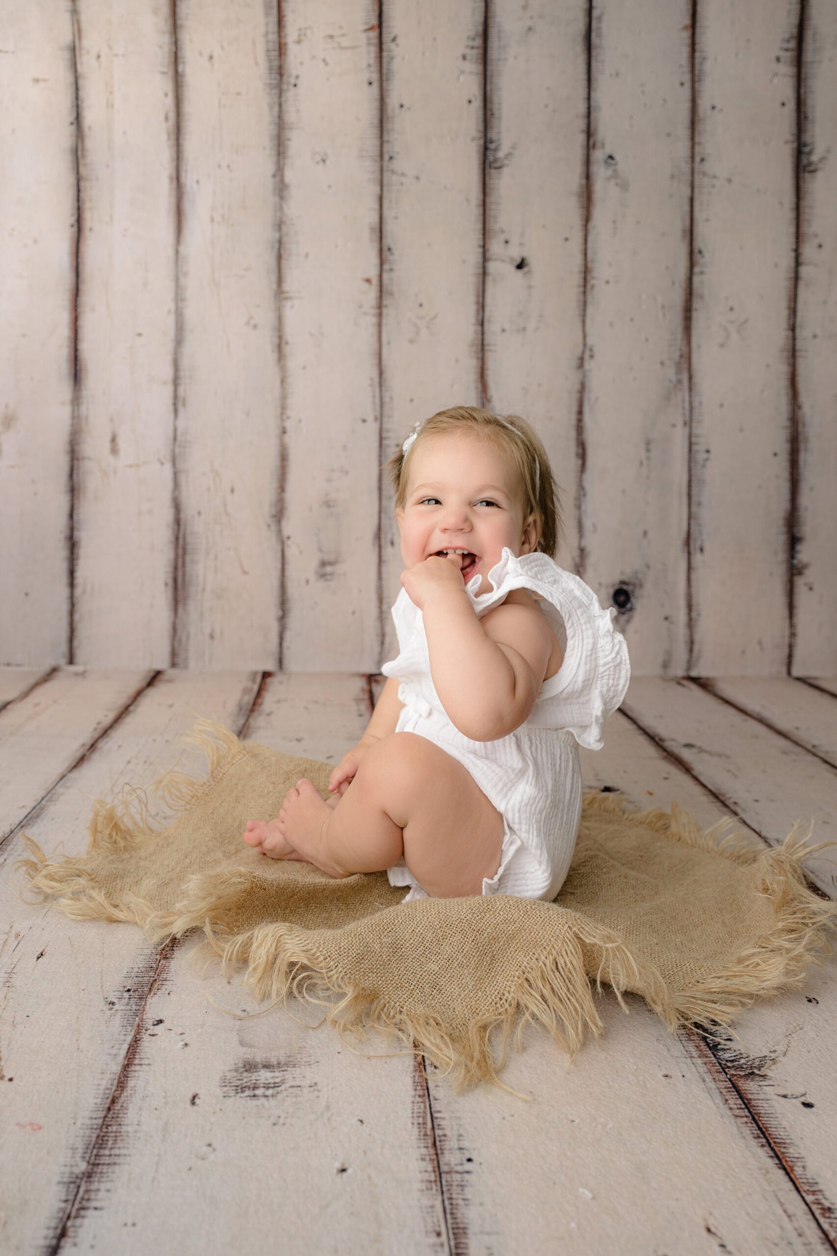 little girl turned to the side with her finger in her mouth dressed in a white romper sitting on burlap