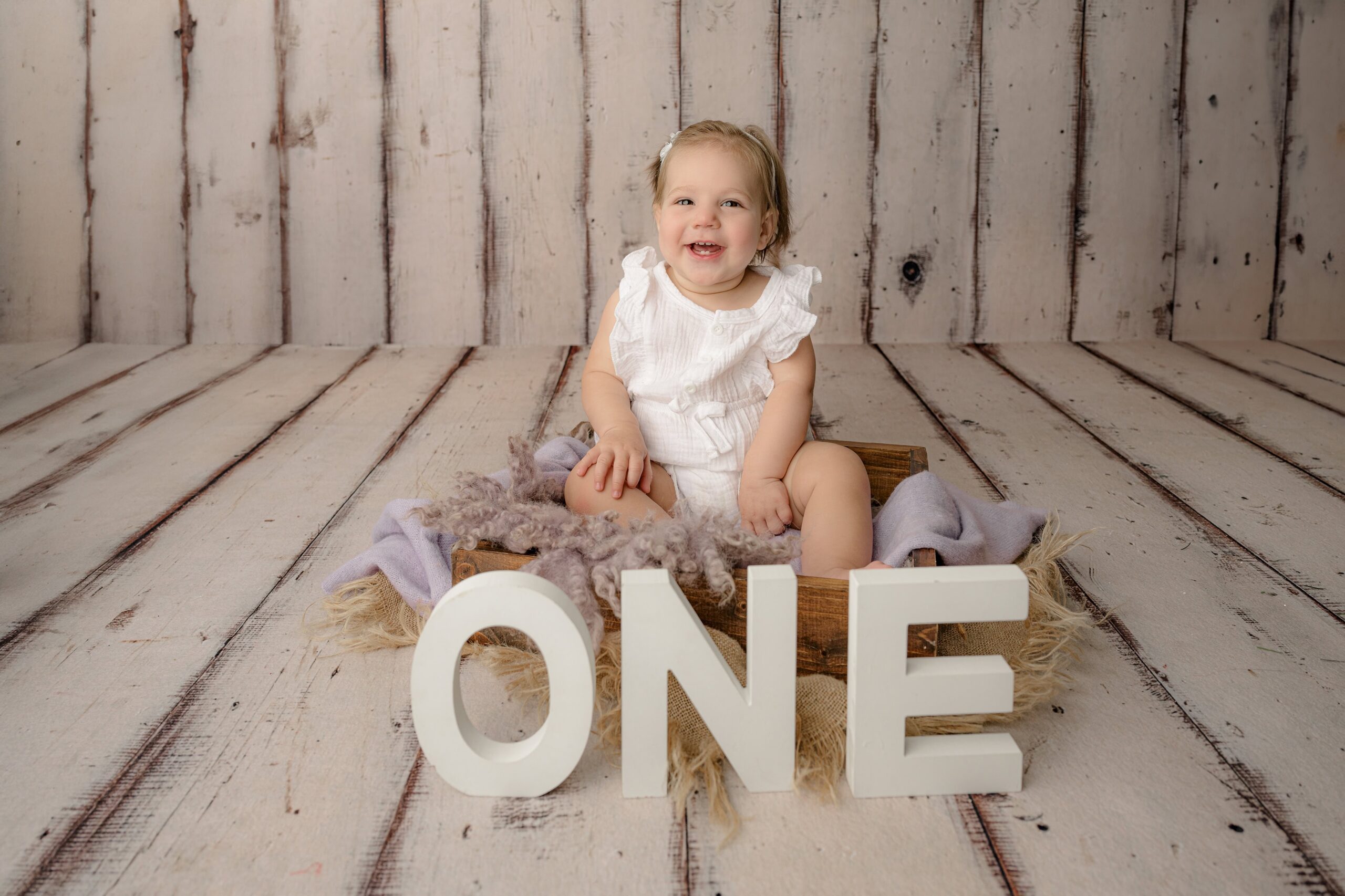 little girl dressed in a white romper sitting on purple cloth in a crate with the letters ONE in front of her