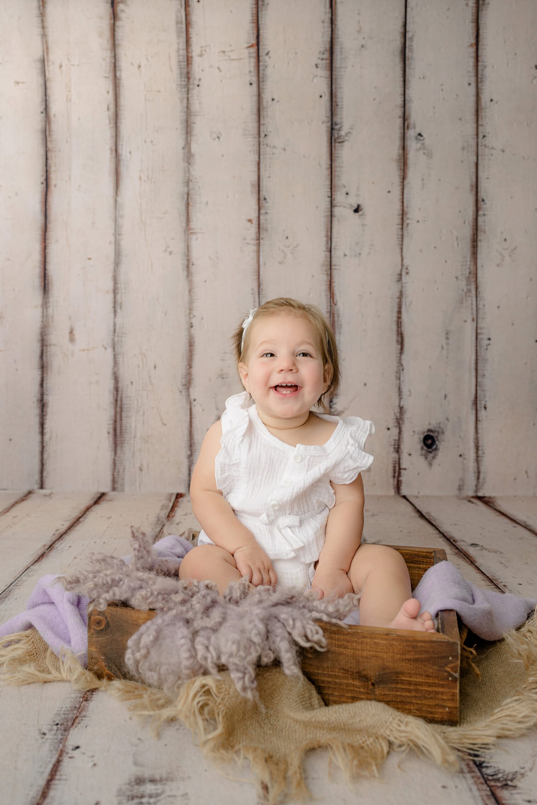 a laughing little girl dressed in a white romper sitting on purple cloth in a crate
