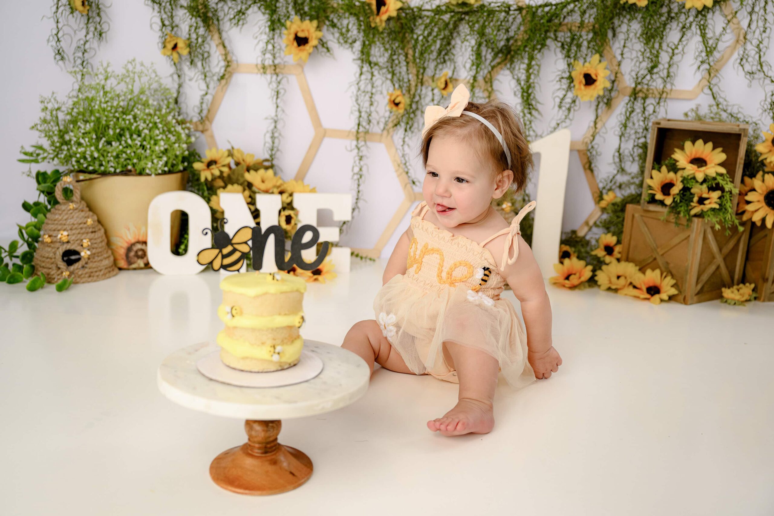 Little girl sits in her peach ONE embroidered romper looking at her cake with her tongue out in a charlotte area nc studio session