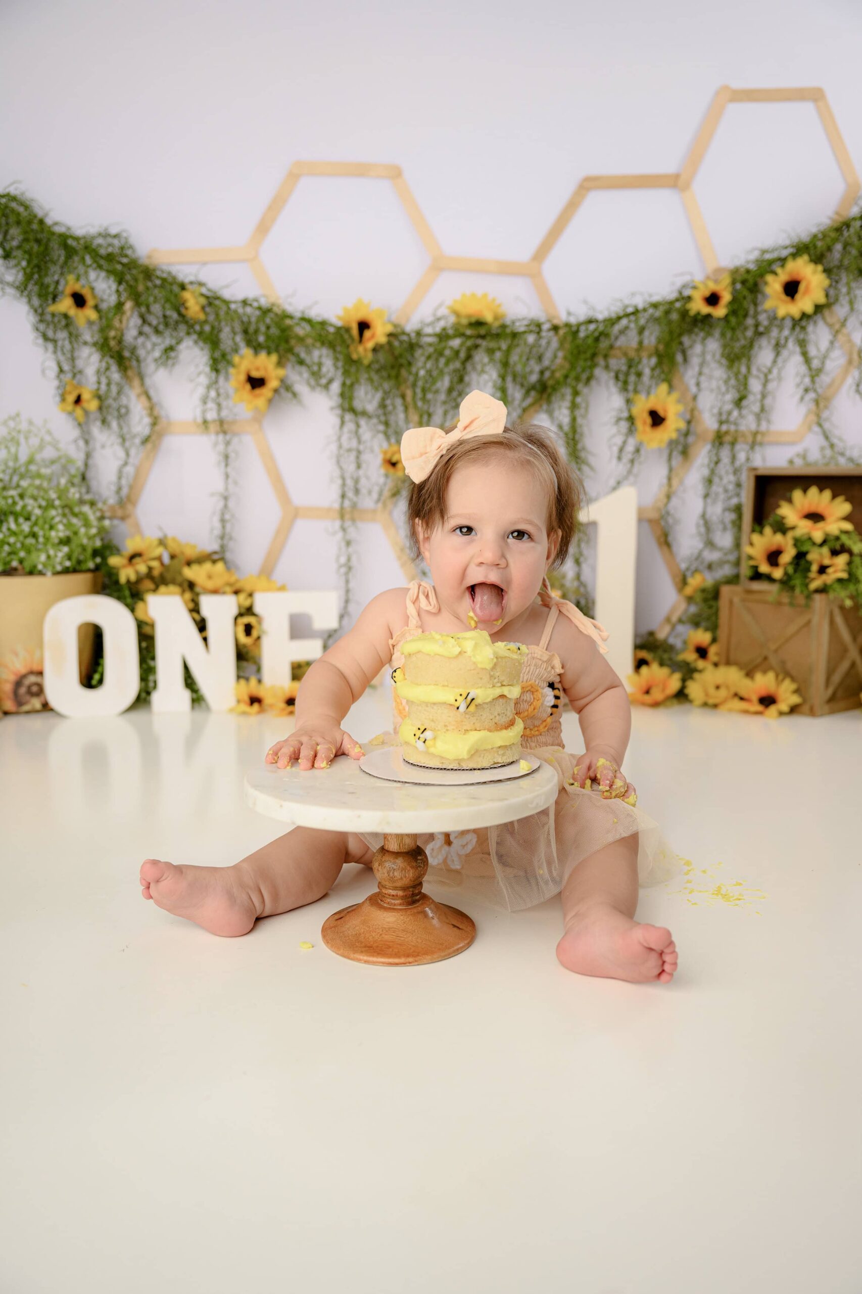 Little girl bending over her multilayered cake taking a big bite out of it during her happy bee day cake smash session